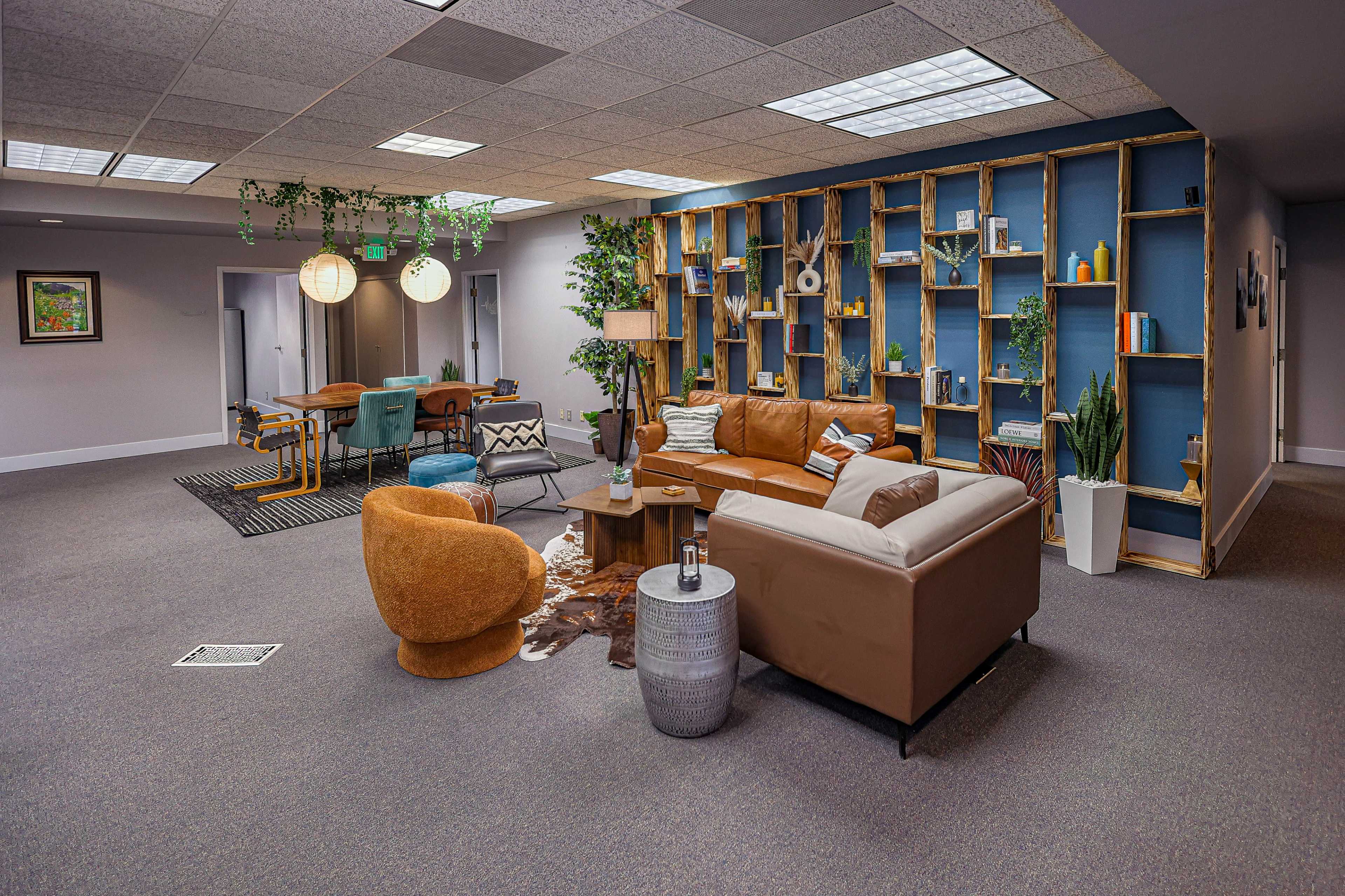 The image shows a modern office space featuring a lounge area with leather sofas, a wooden coffee table, and a decorative bookshelf, alongside a dining area with colorful chairs and a patterned rug.