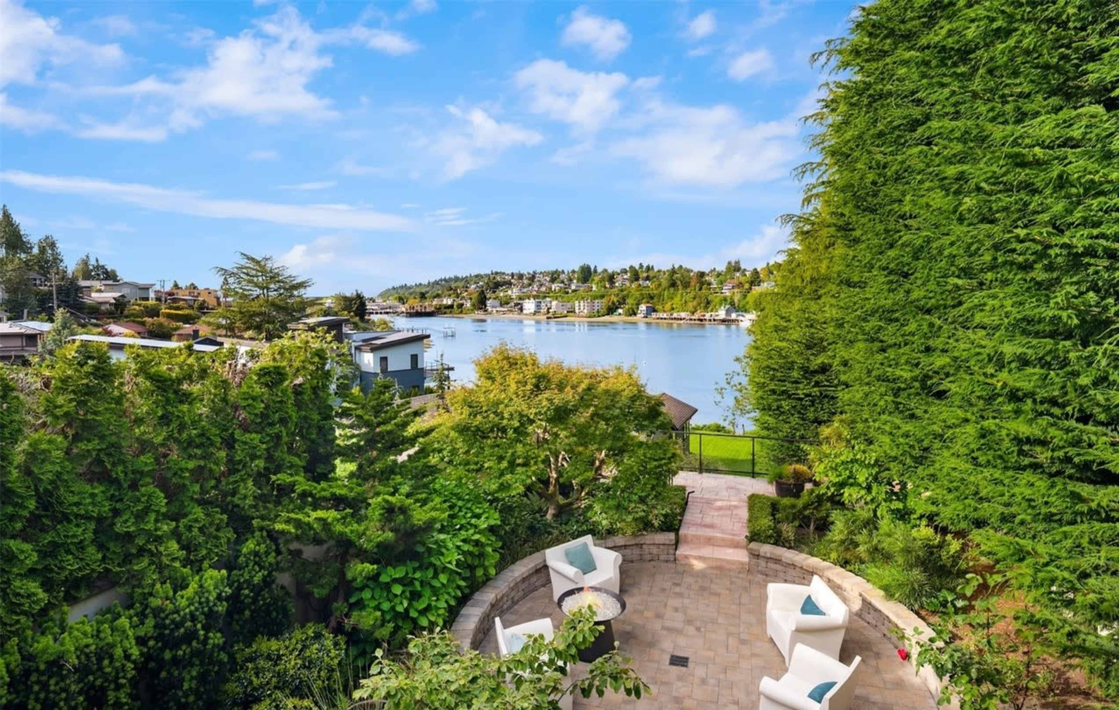 A waterfront view from a landscaped terrace featuring white chairs and greenery overlooking a calm body of water with houses in the distance.
