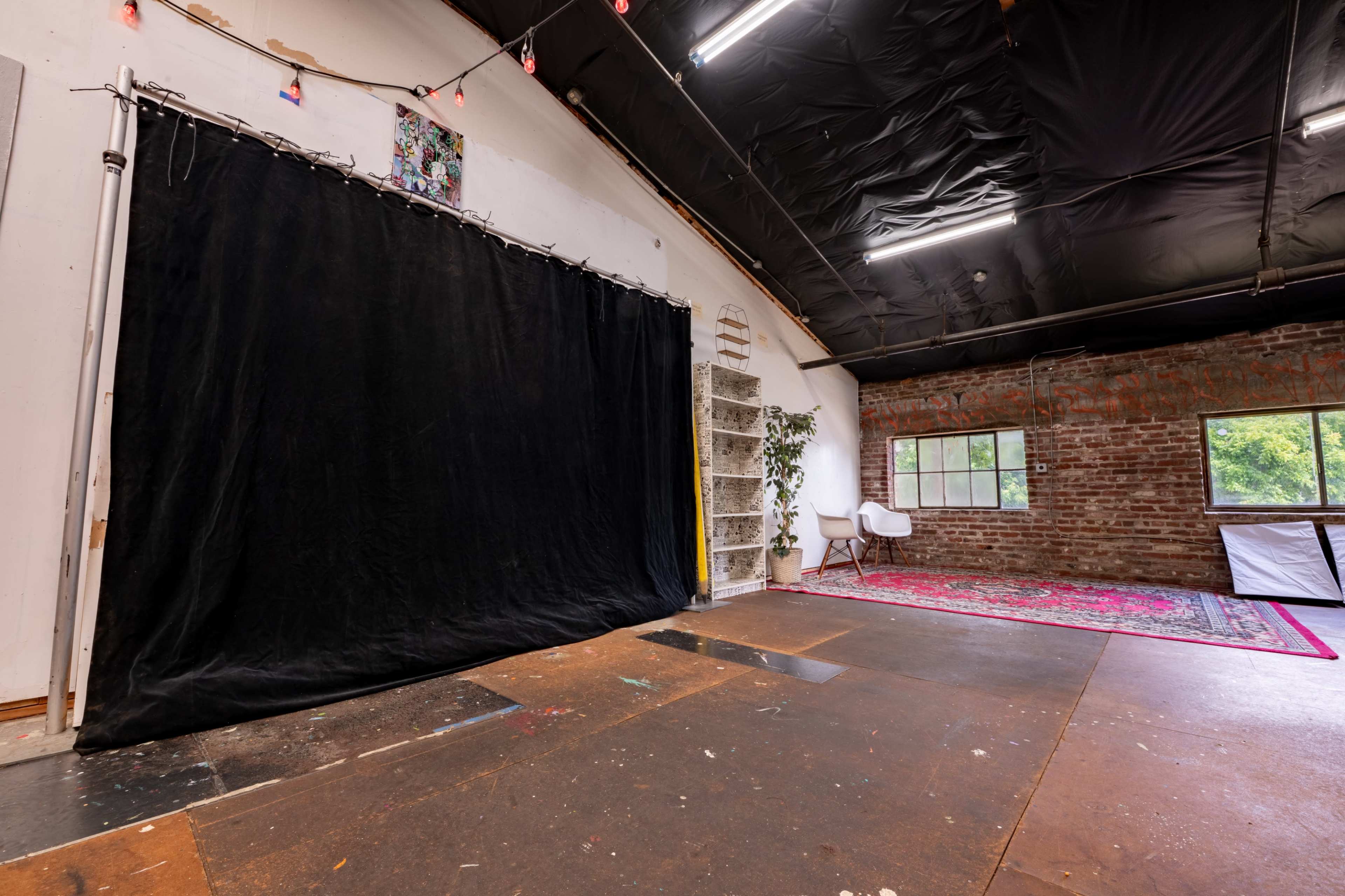 The image shows an art studio with a black backdrop, a brick wall, and a white chair placed on a patterned rug.