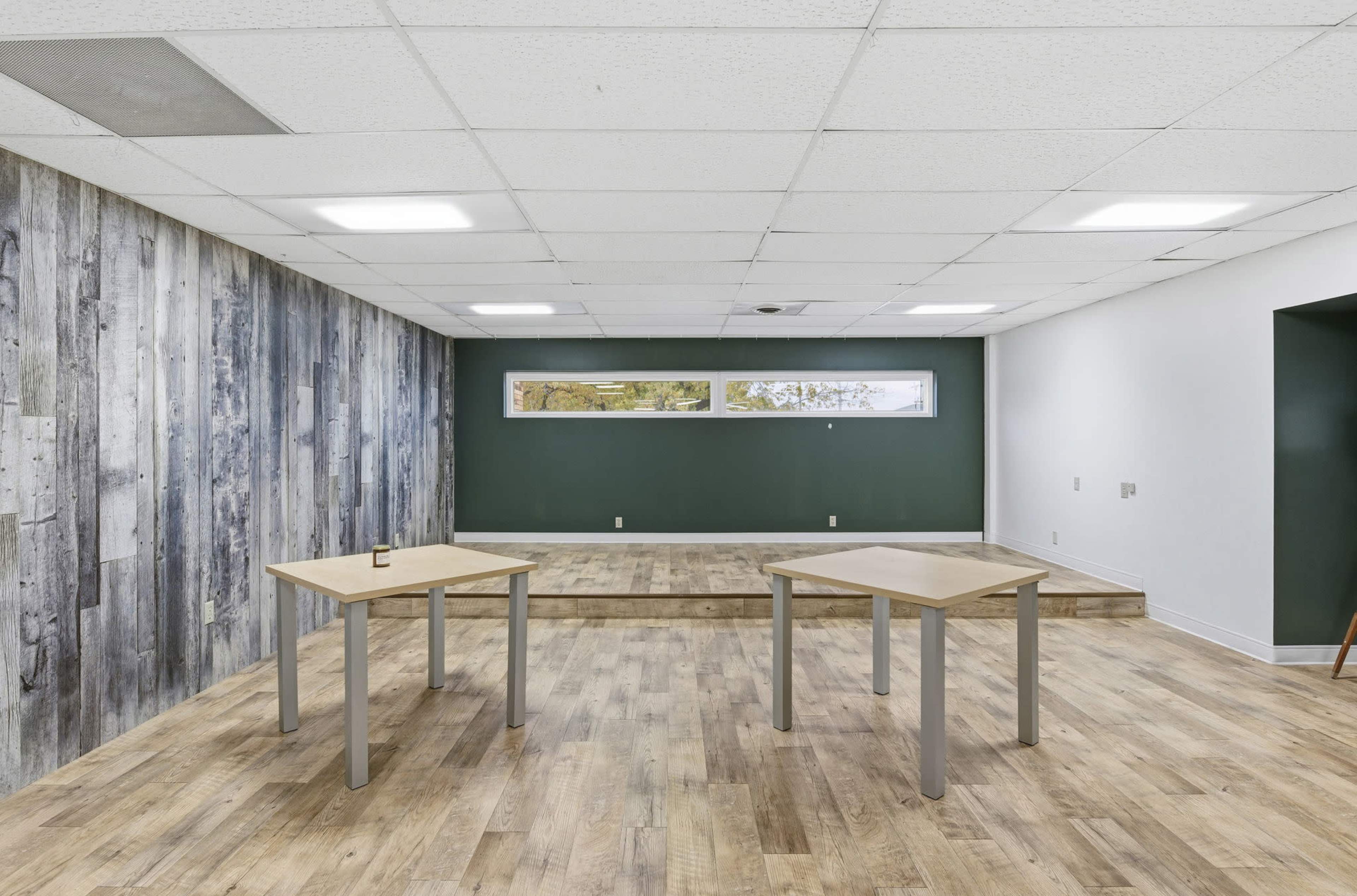 The image shows an empty room with wooden flooring, two simple tables, and a raised platform against a green wall with a large window.