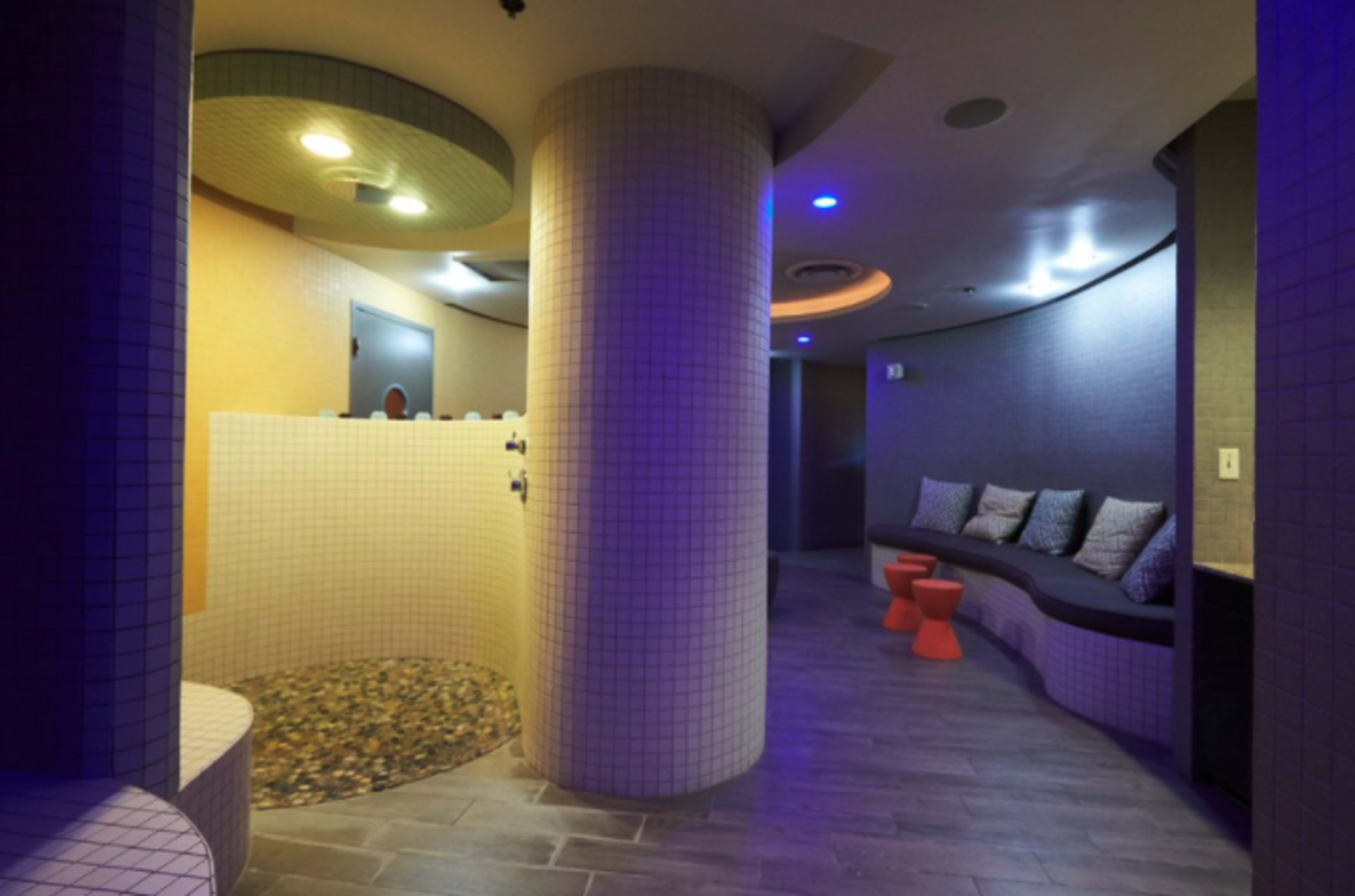 The image shows a modern spa interior featuring curved walls, a seating area with cushions, and a tiled floor with a pebble section.
