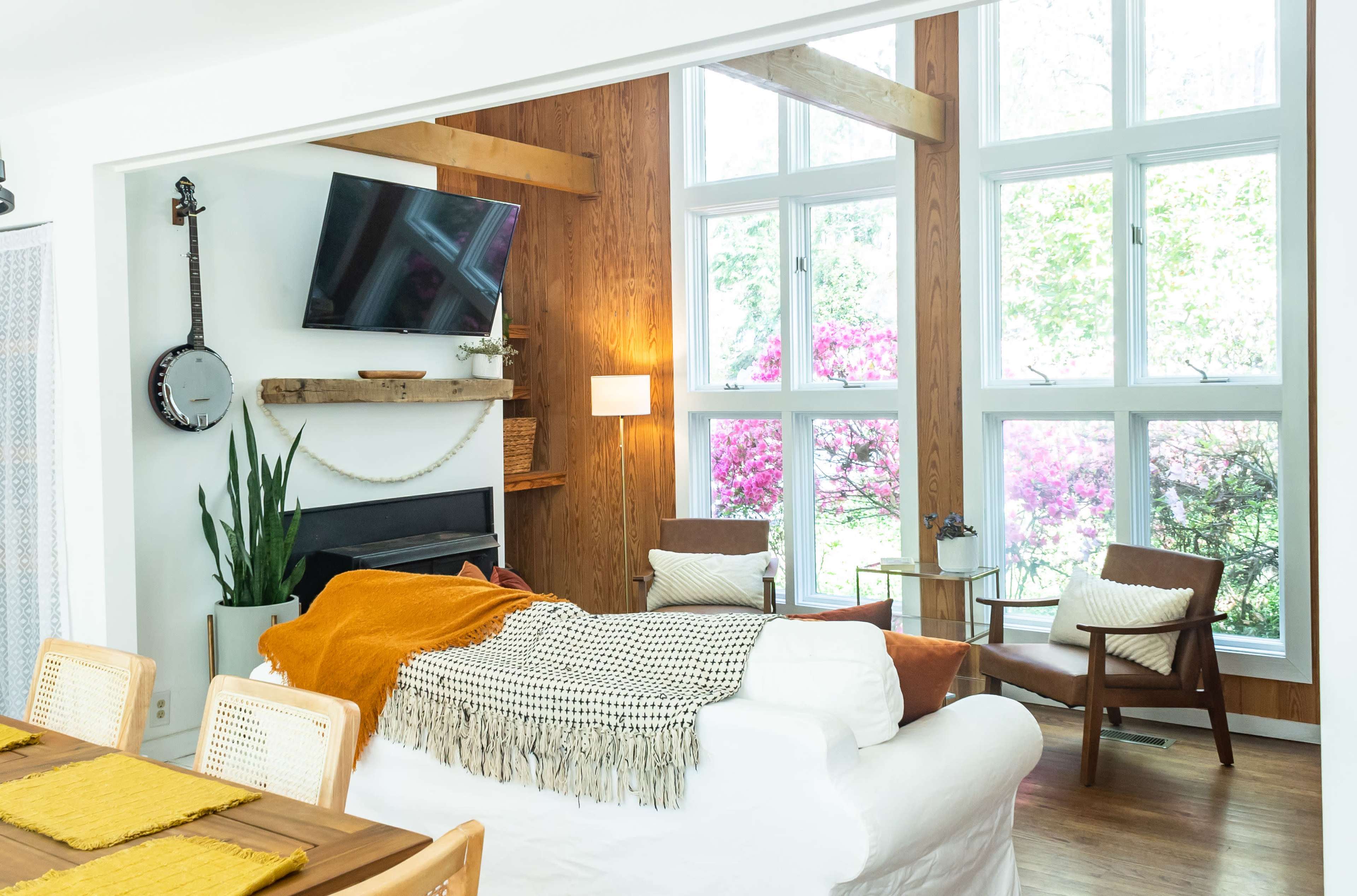 A bright living room features a white sofa adorned with a blanket, a wooden dining table, and large windows that overlook blooming flowers outside.