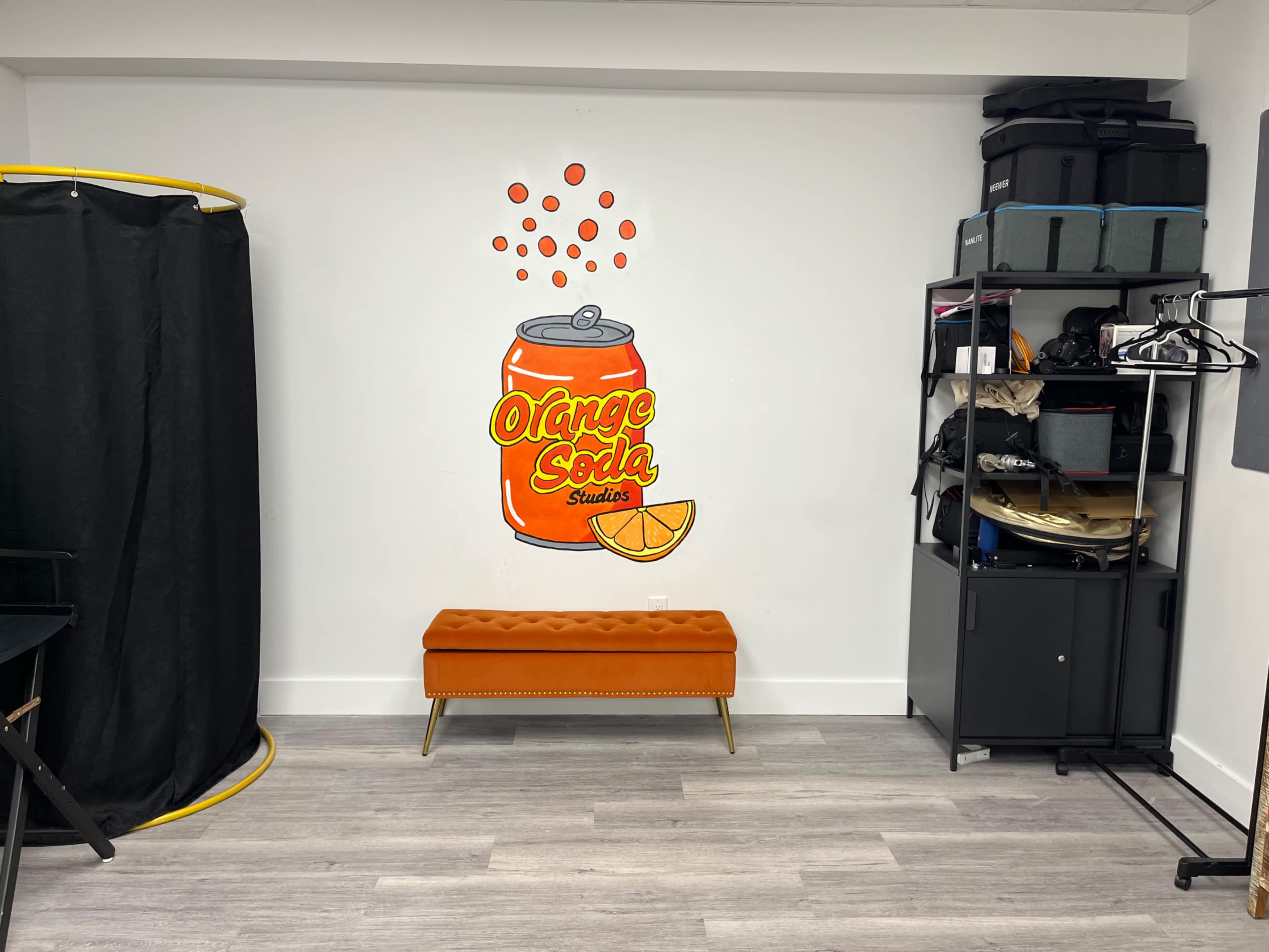 The image shows a small room with an orange bench in front of a wall featuring a mural of an orange soda can, along with black storage units and a curtain in the corner.