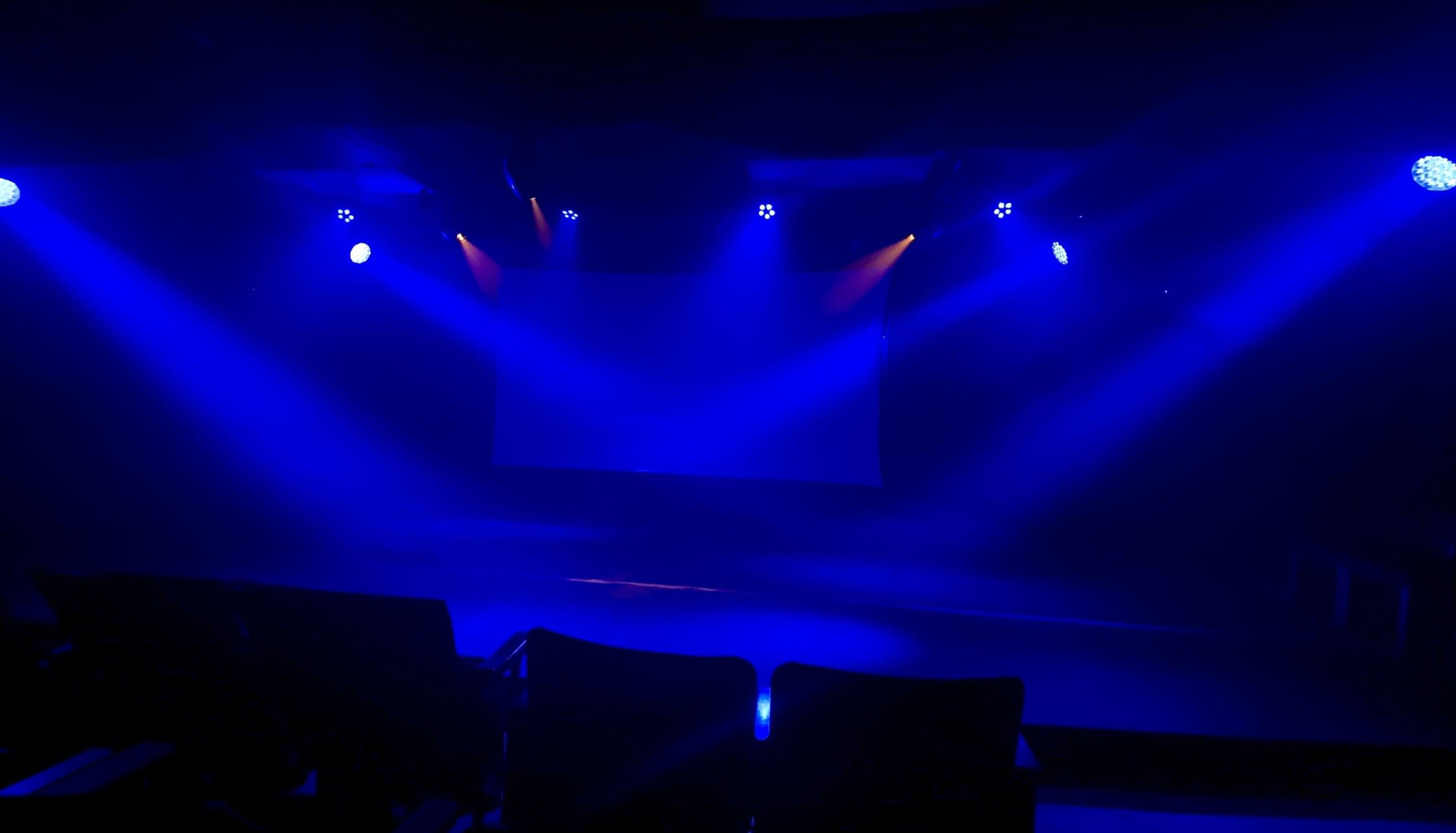 A dimly lit stage with blue lighting and empty chairs facing a blank projection screen.