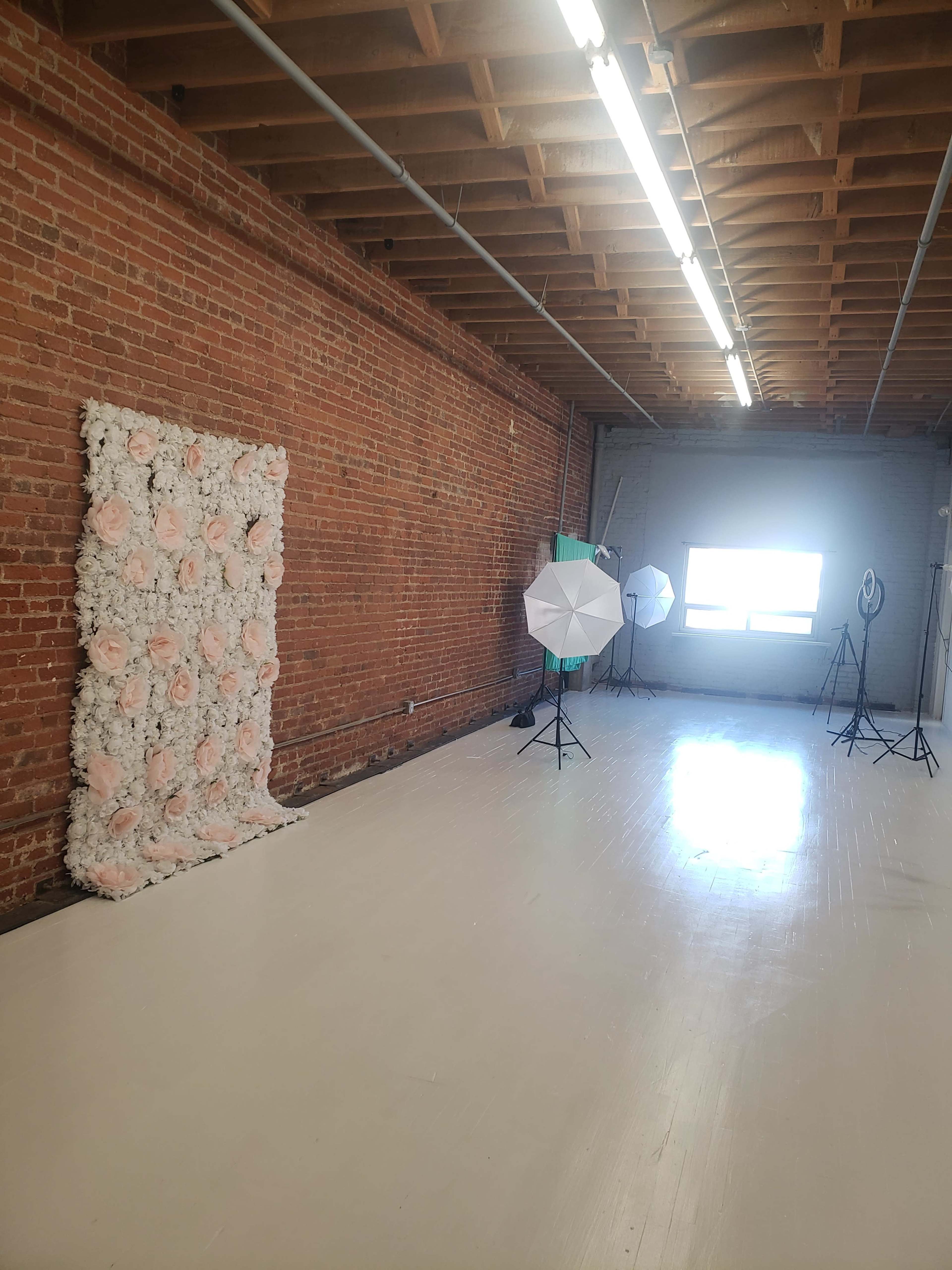 The image shows a spacious photography studio with a floral backdrop, lighting equipment, and exposed brick walls.