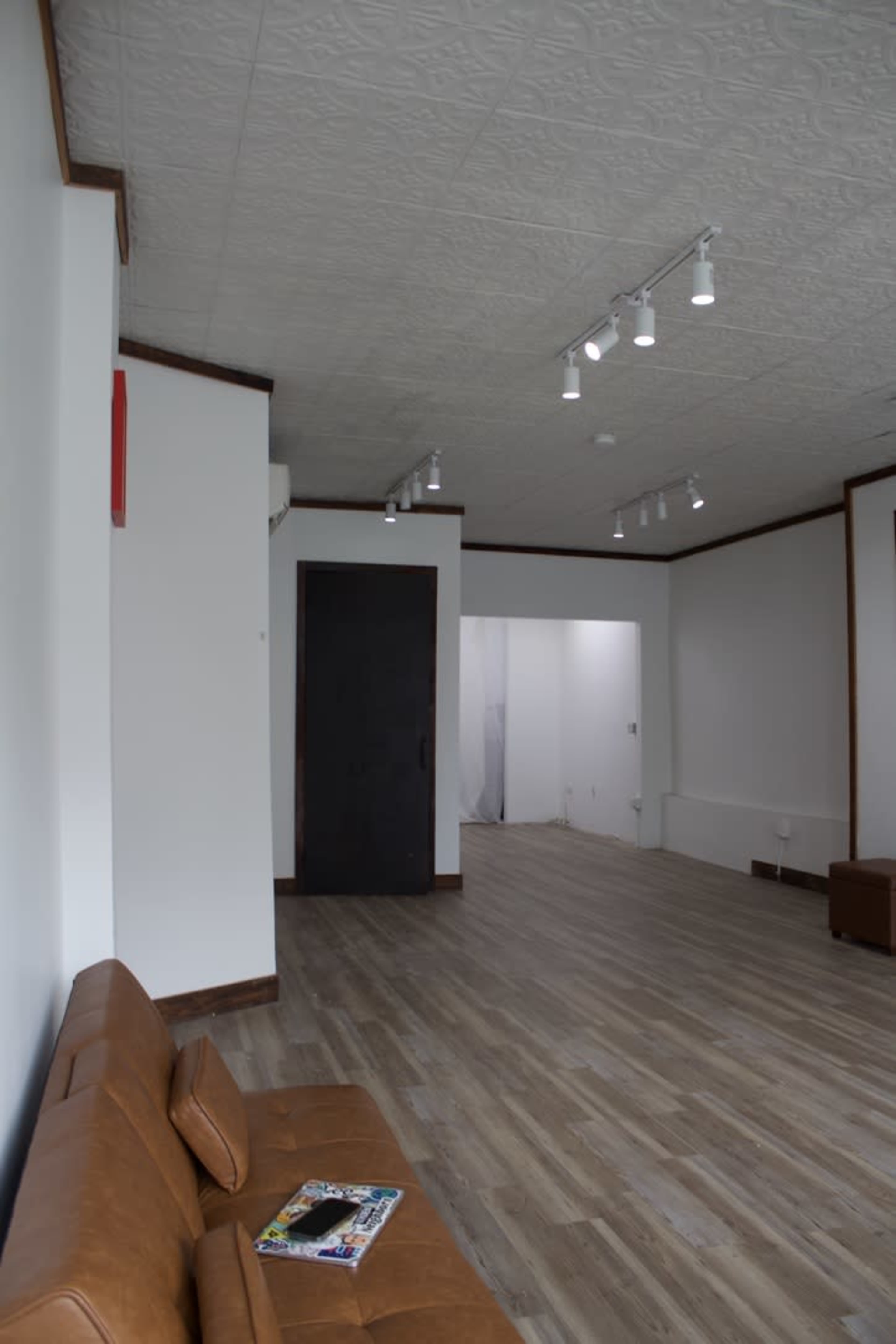 The image shows a minimalist interior of a room with light wooden flooring, white walls, and a brown leather sofa.