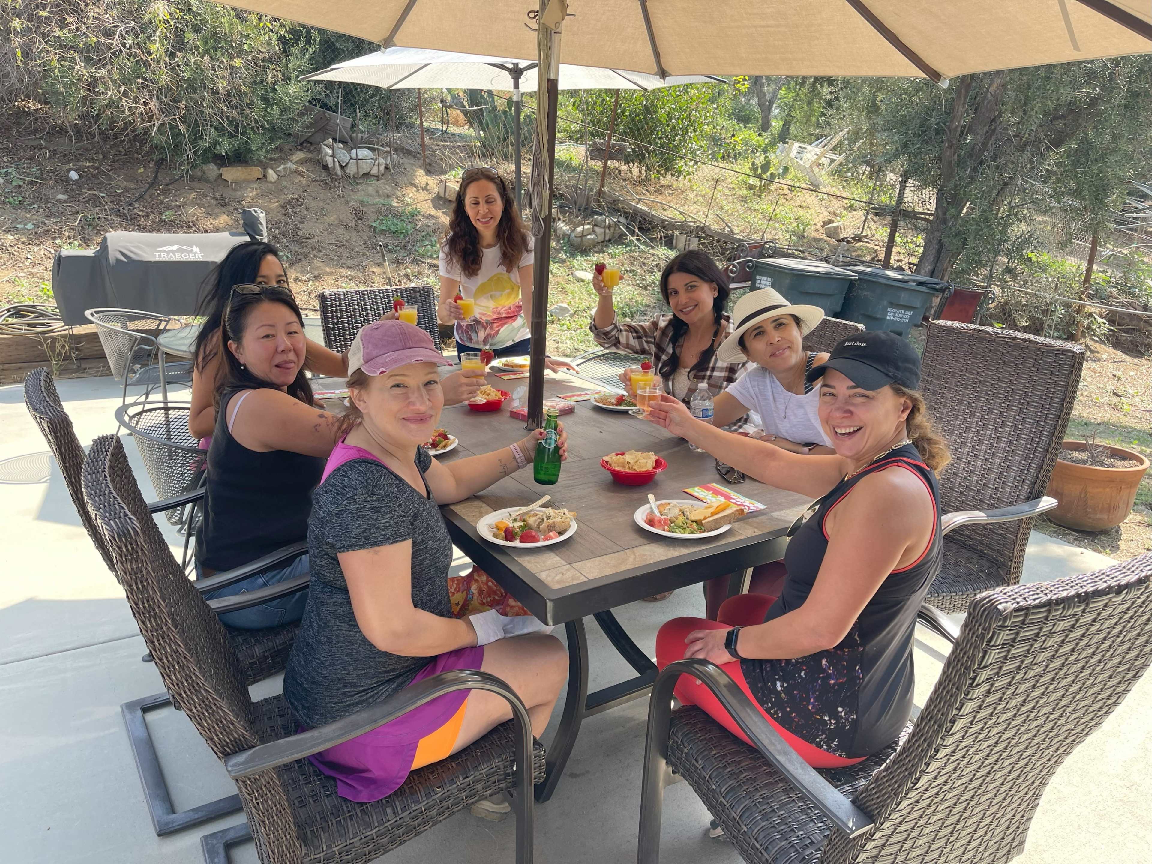 A group of six women sits around a table under a large umbrella, sharing a meal outdoors.