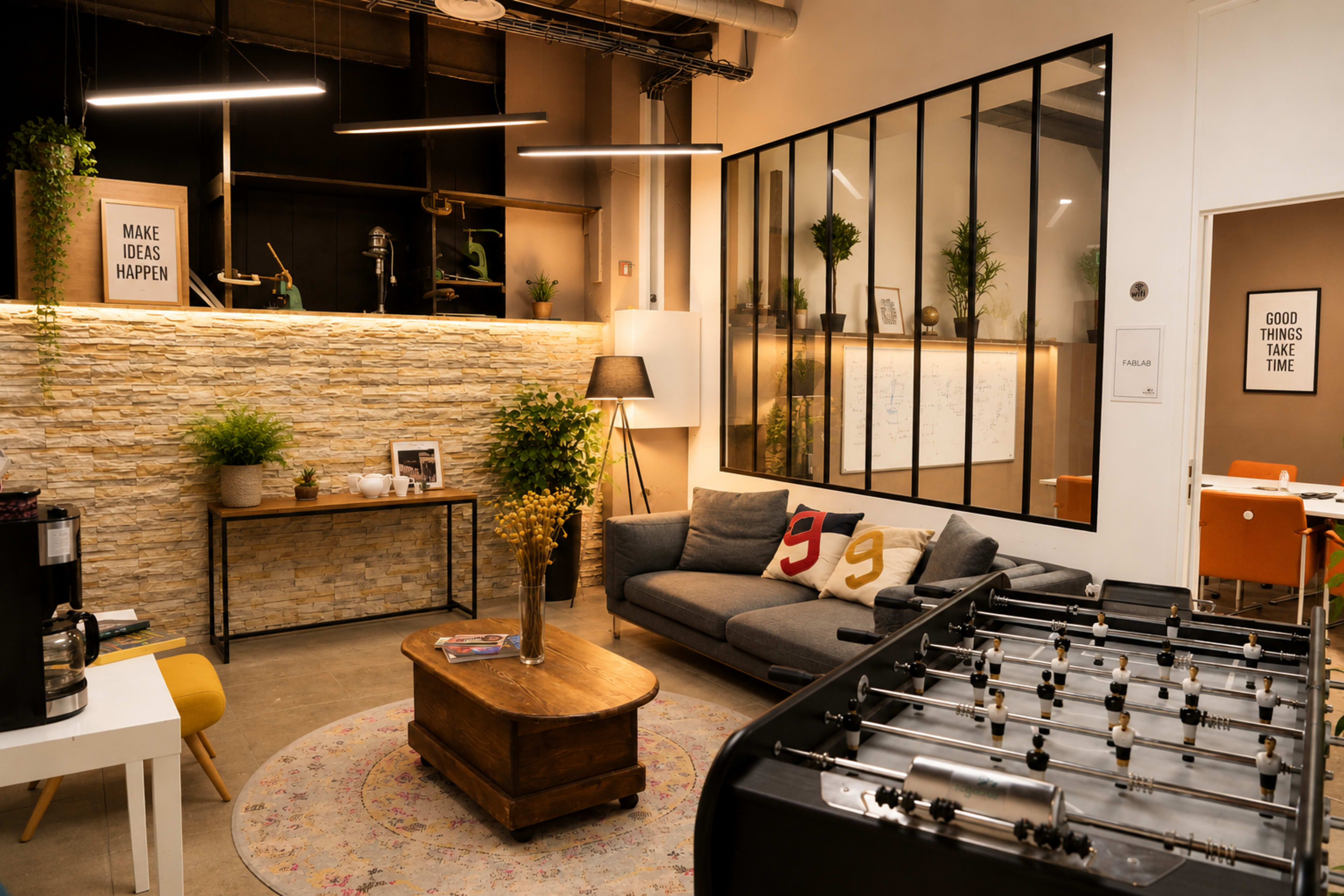 The image shows a modern office space featuring a cozy seating area, a foosball table, and a decorative stone wall with plants and framed motivational quotes.