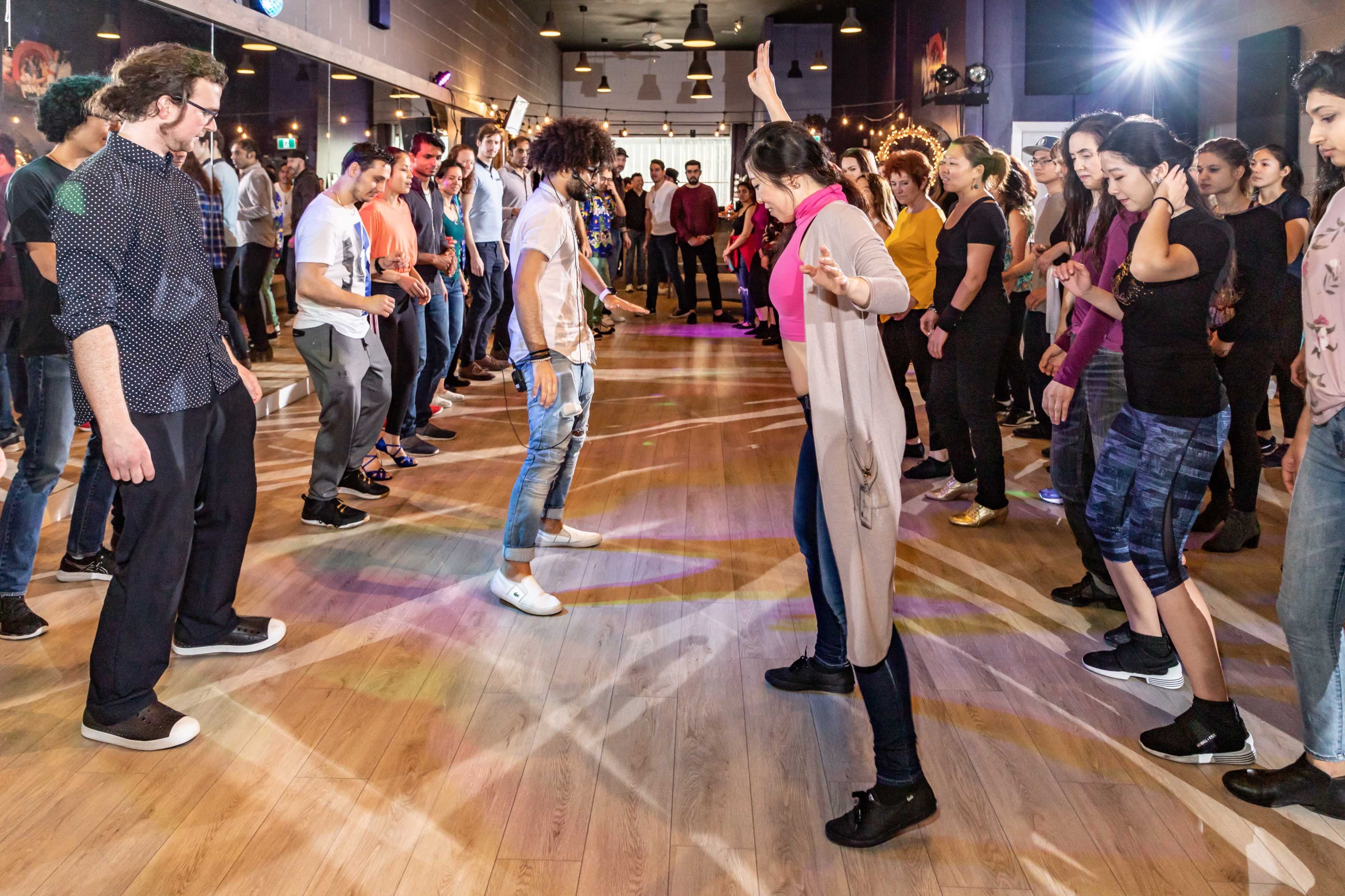 A large group of people are gathered in a dance studio, participating in a dance class with one instructor leading in the center.