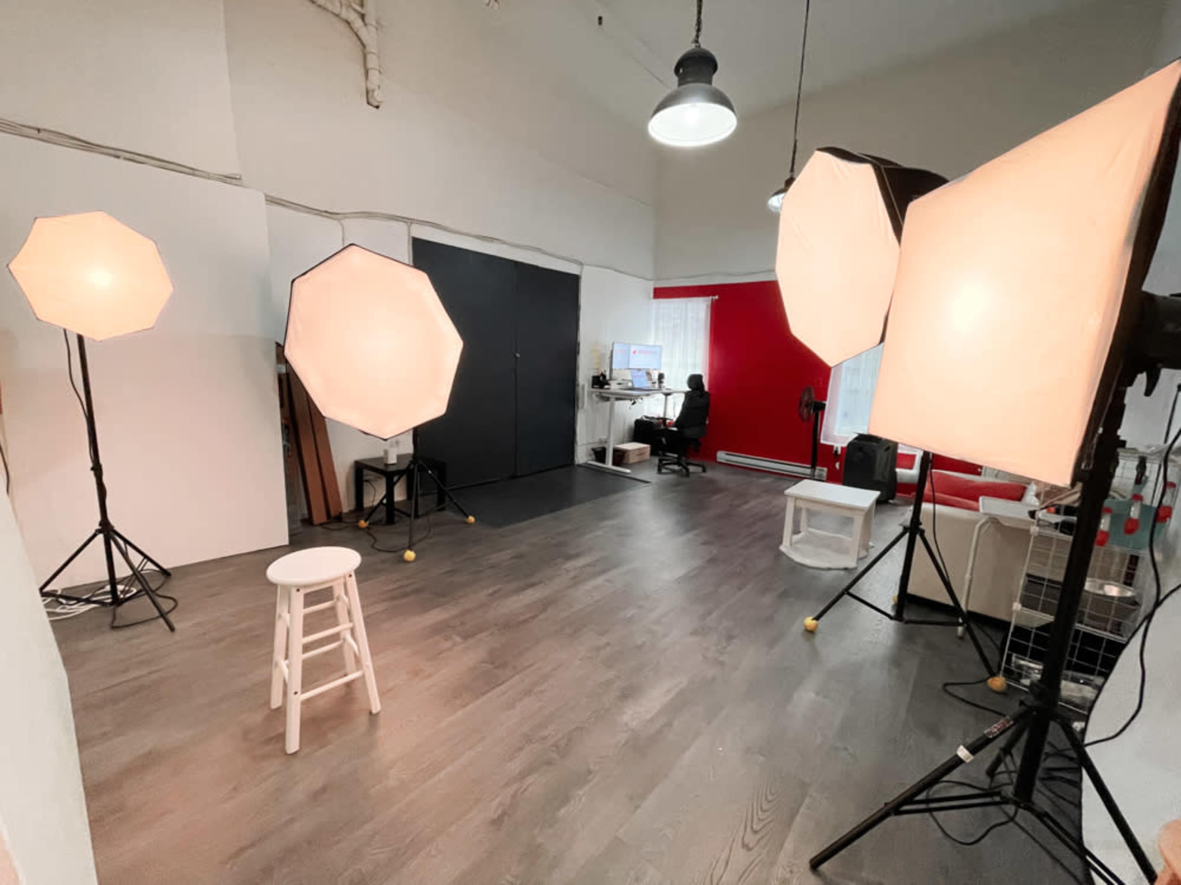 A photography studio features four large softbox lights, a white stool, and a workspace with a computer against a red accent wall.