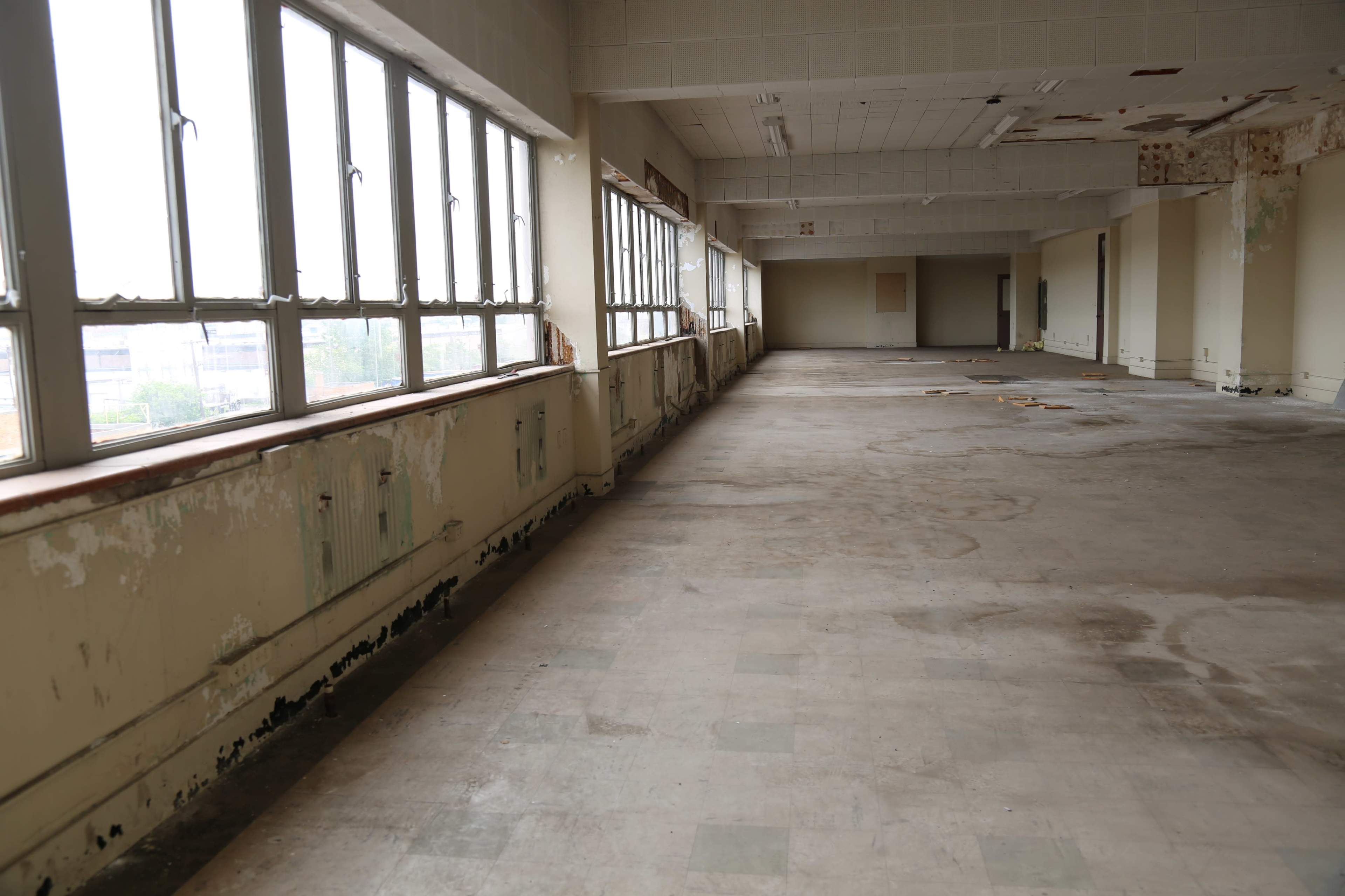 A large, empty room with worn flooring and windows along one side, allowing natural light to enter.