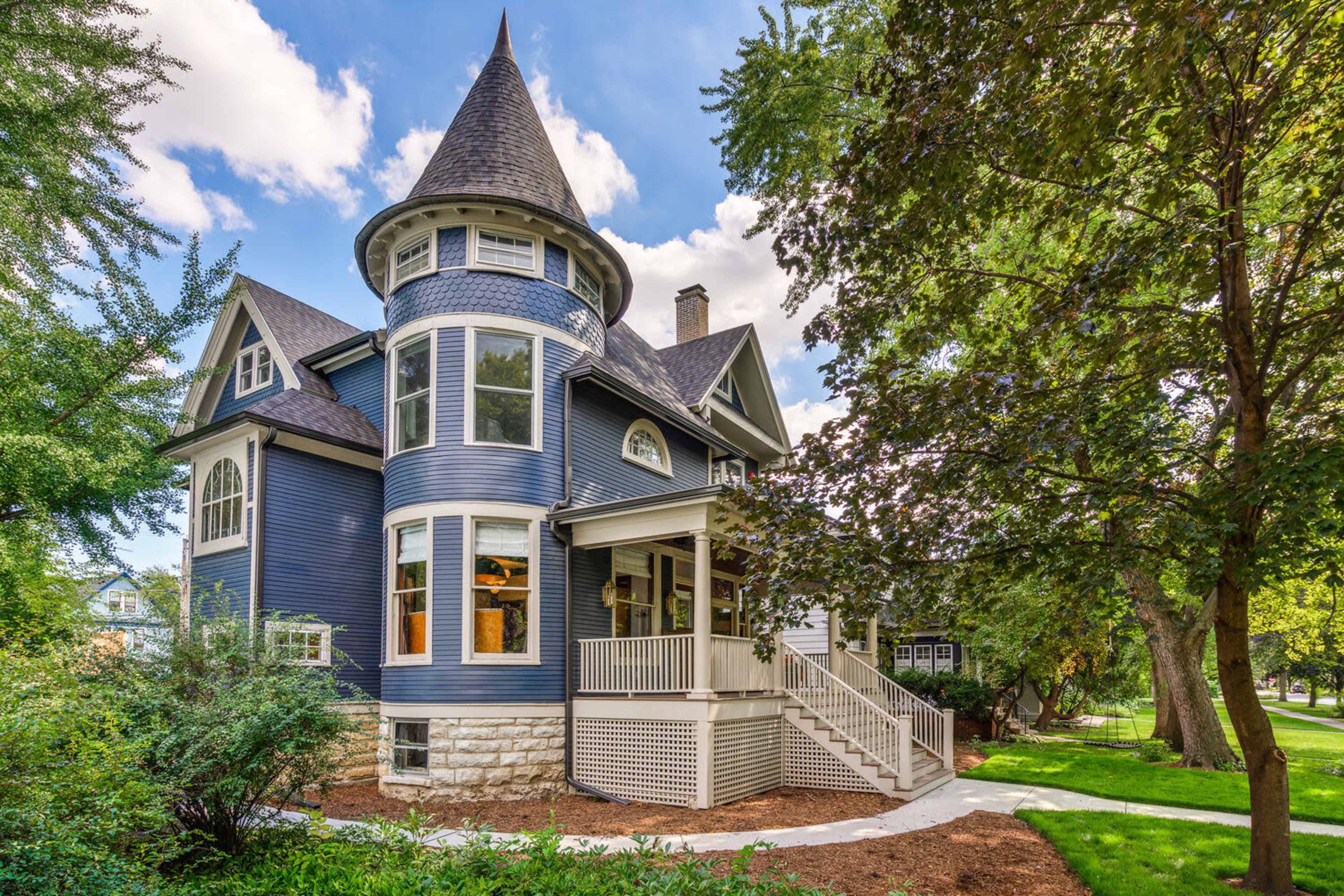 A large blue Victorian-style house with a turret and a wraparound porch, surrounded by green trees and landscaping.