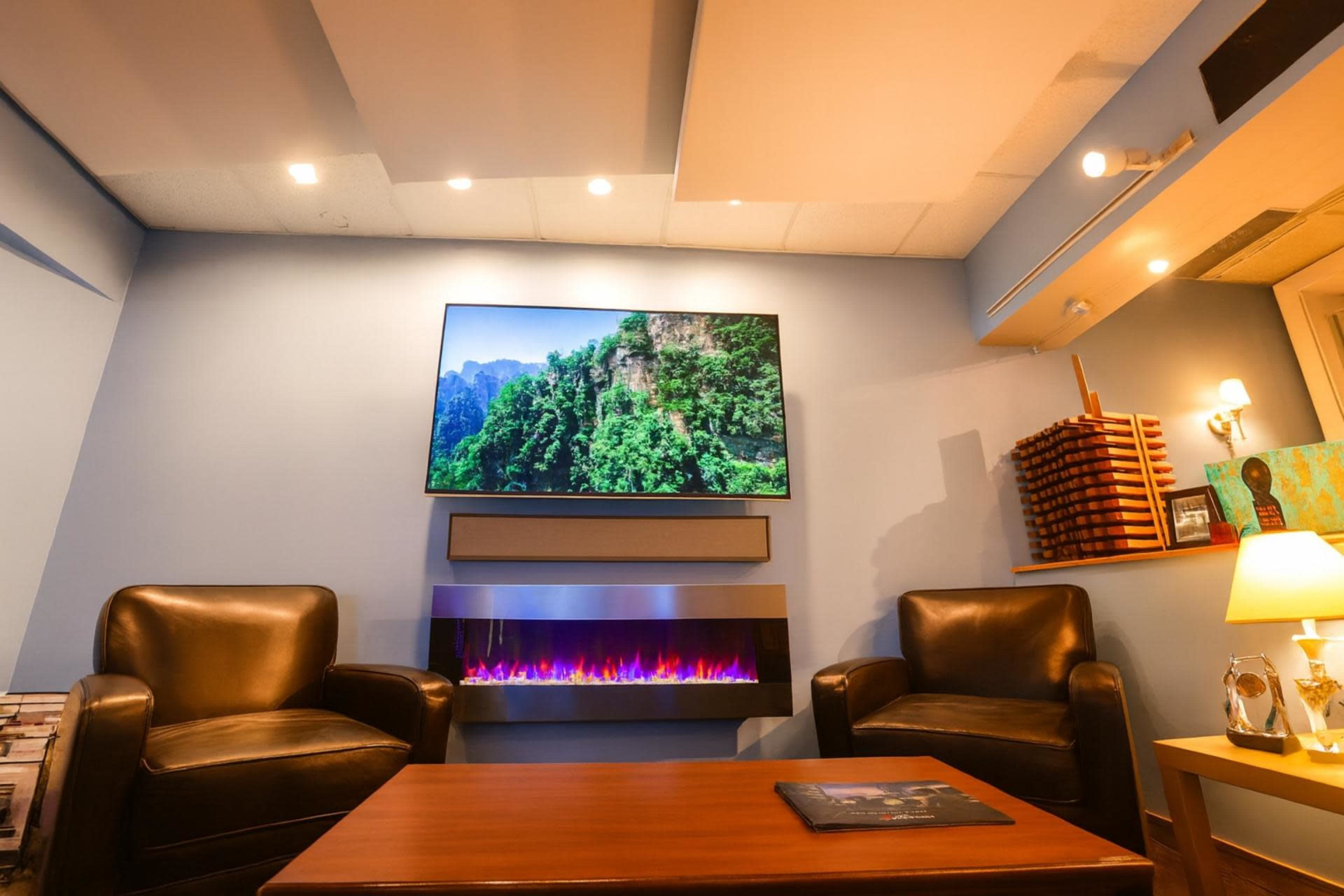 The image shows a cozy room with two leather armchairs facing a modern electric fireplace and a large mounted television displaying a scenic mountain landscape.