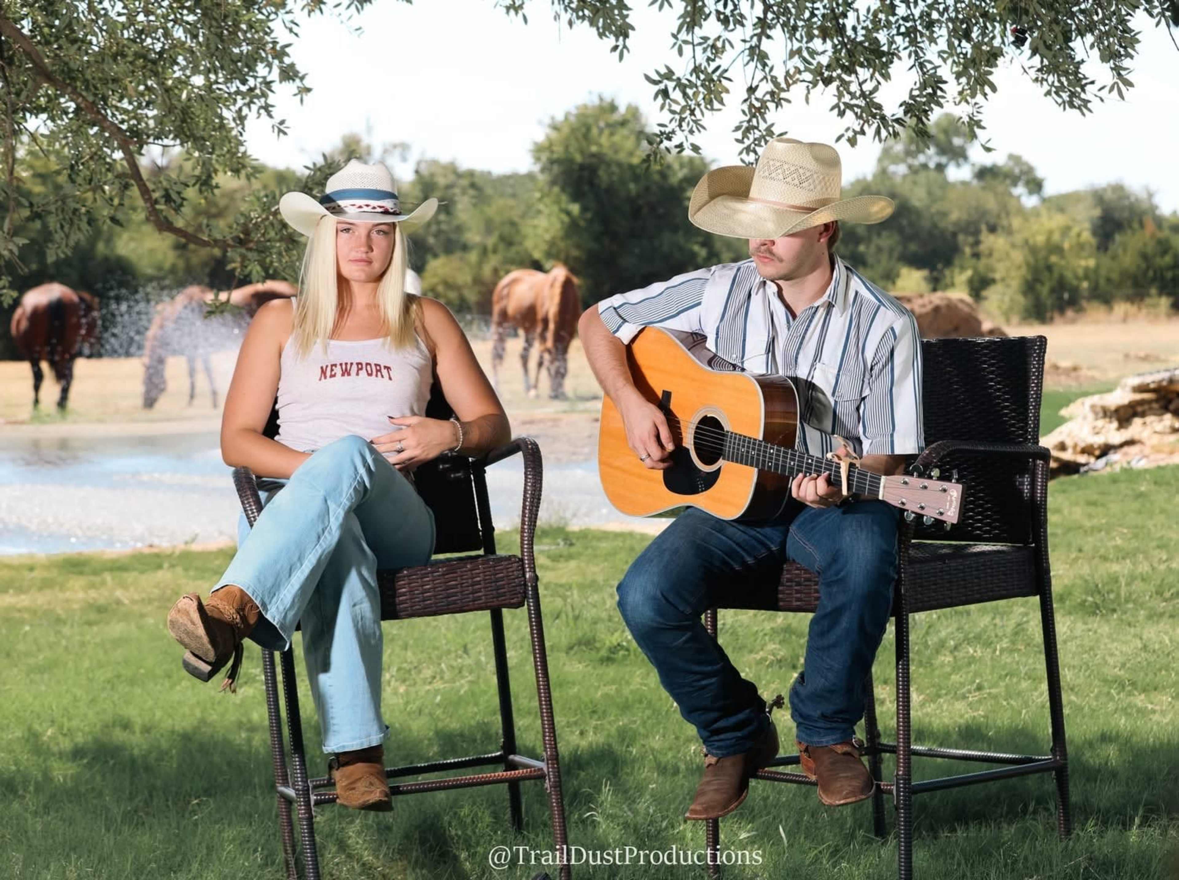 A woman sits on a chair next to a man playing guitar, both in cowboy attire, with horses grazing in the background near a pond.