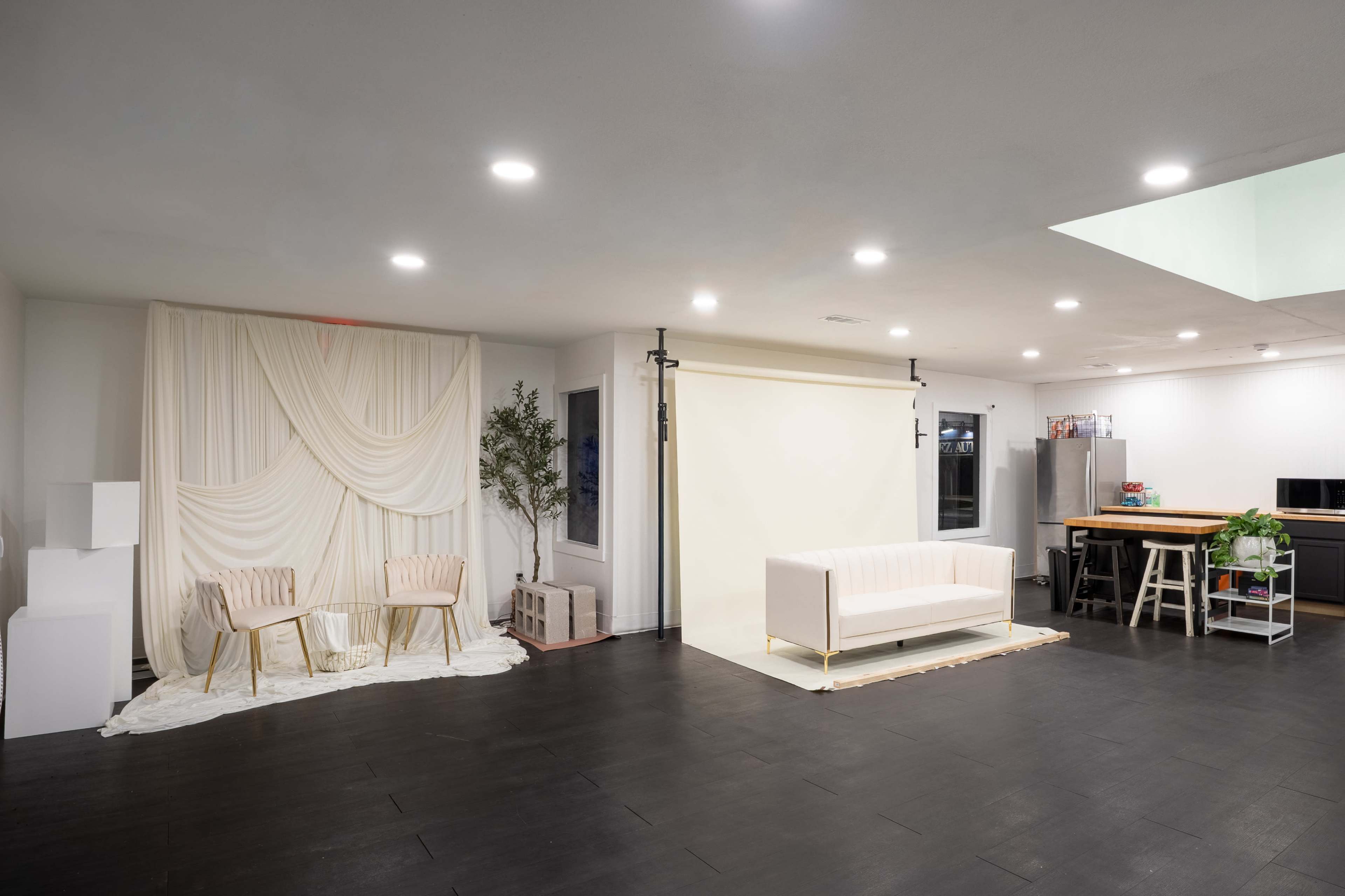 The image shows a spacious indoor room with a white backdrop and seating area on one side, and a kitchenette with appliances on the other.