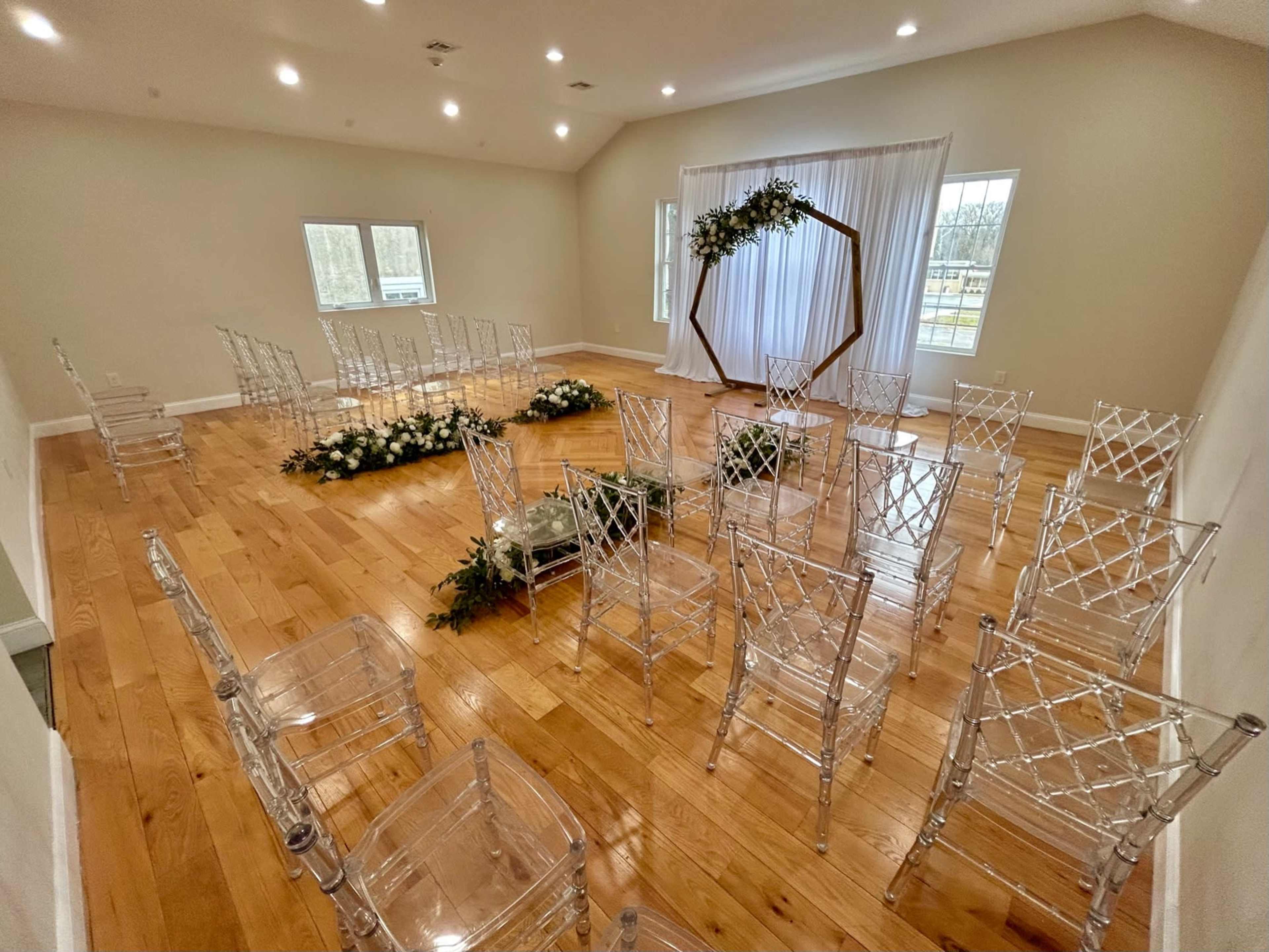 A room is set up for a ceremony, featuring transparent chairs arranged in a circular formation around a decorated altar with floral accents.