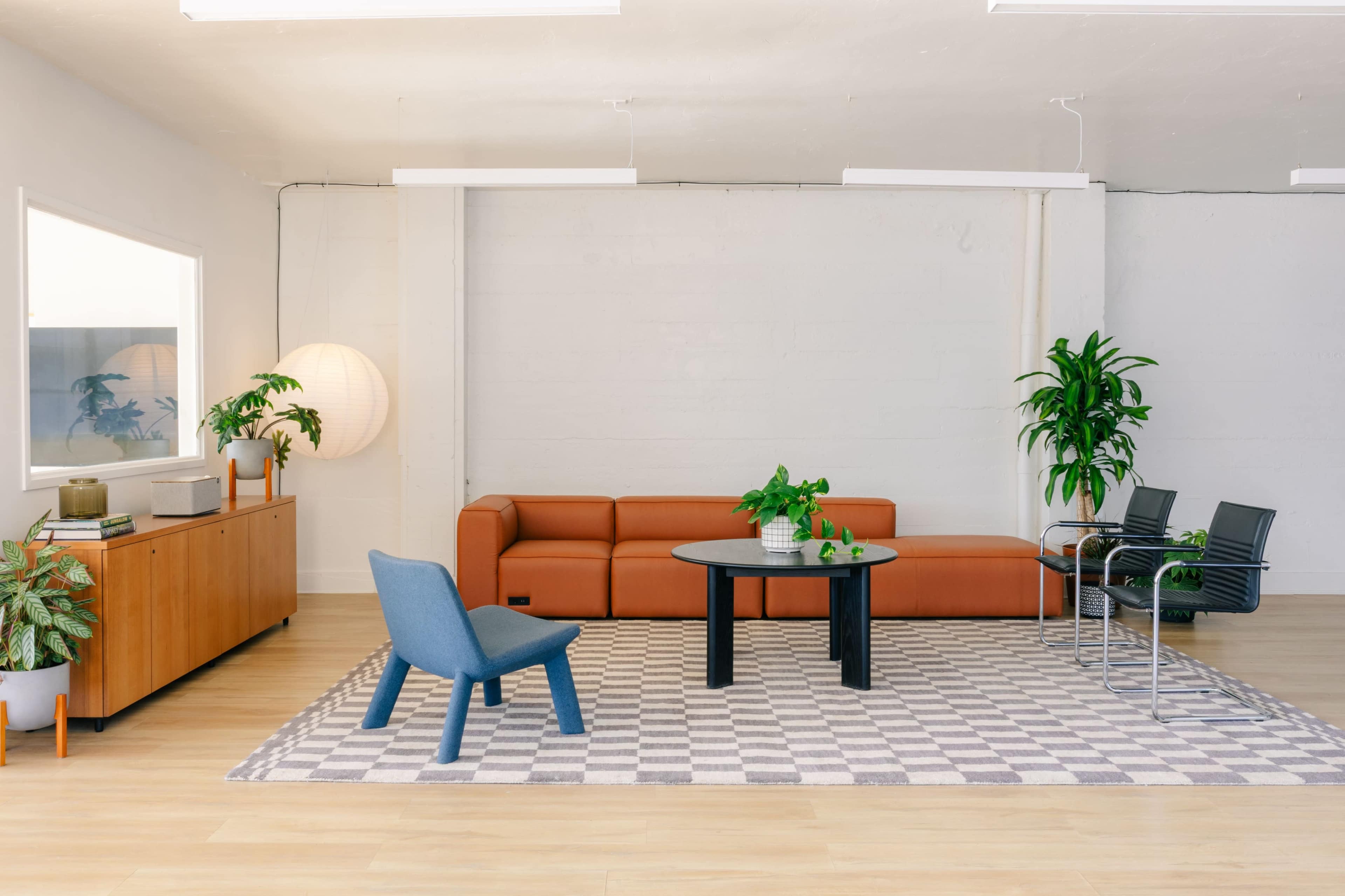 The image features a modern living space with a brown leather sofa, a round black table, stylish chairs, and various indoor plants on a light-colored floor.