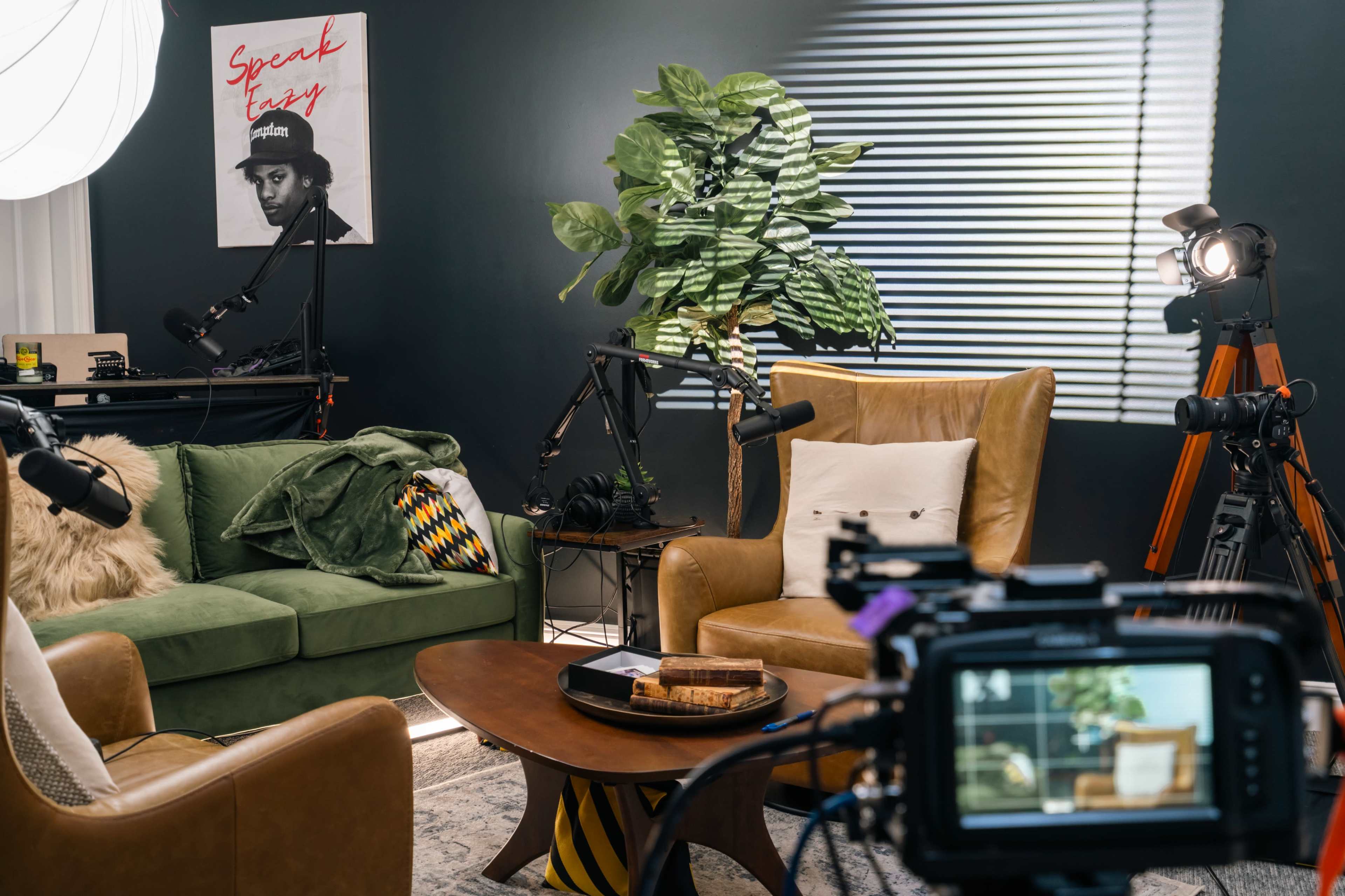 A modern setup for a video recording, featuring two armchairs, a coffee table, and multiple cameras and microphones positioned around a small living space with green walls and a large plant.