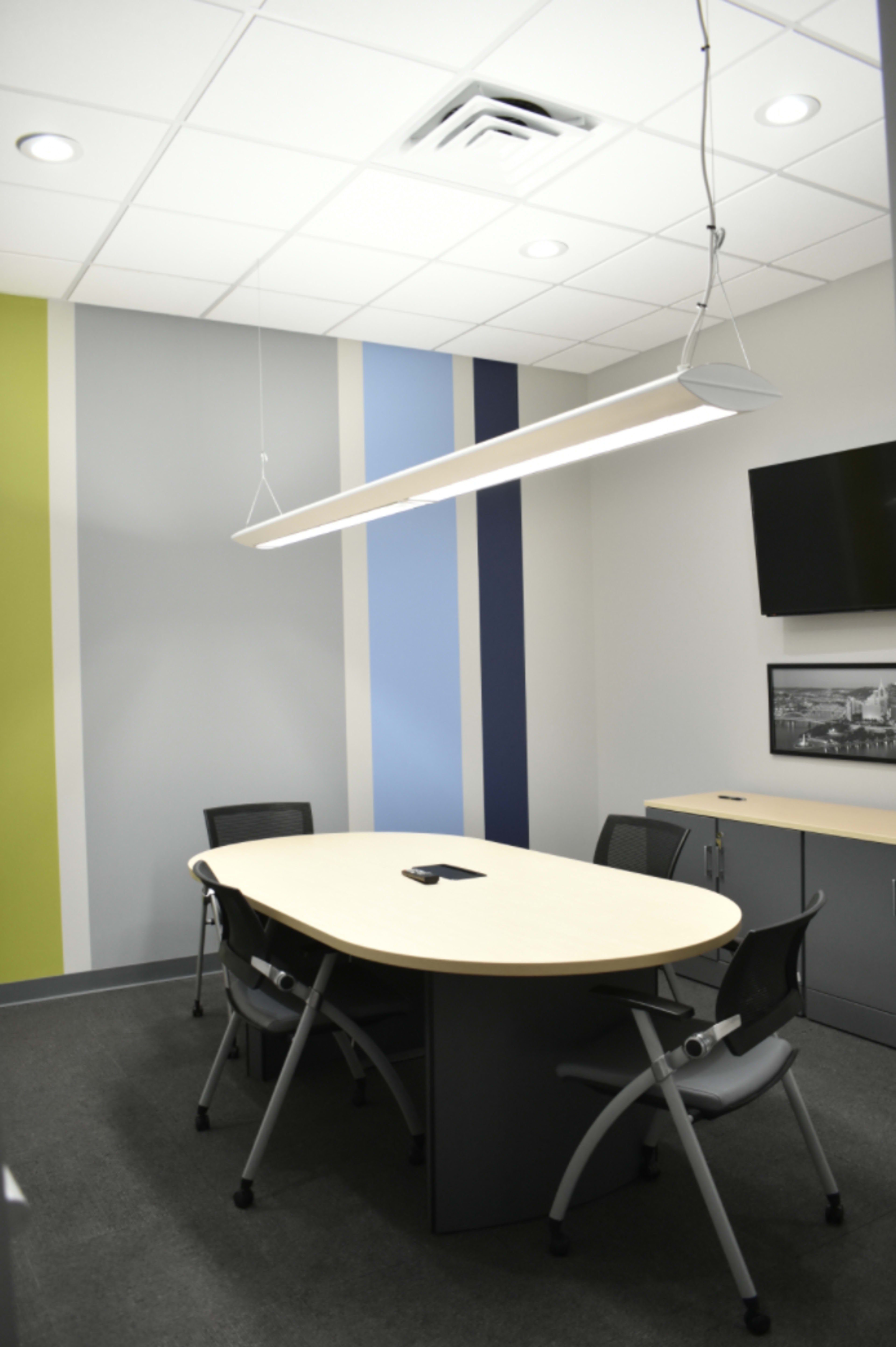 A conference room with a large oval table and four chairs, featuring gray and colored striped walls and a wall-mounted television.