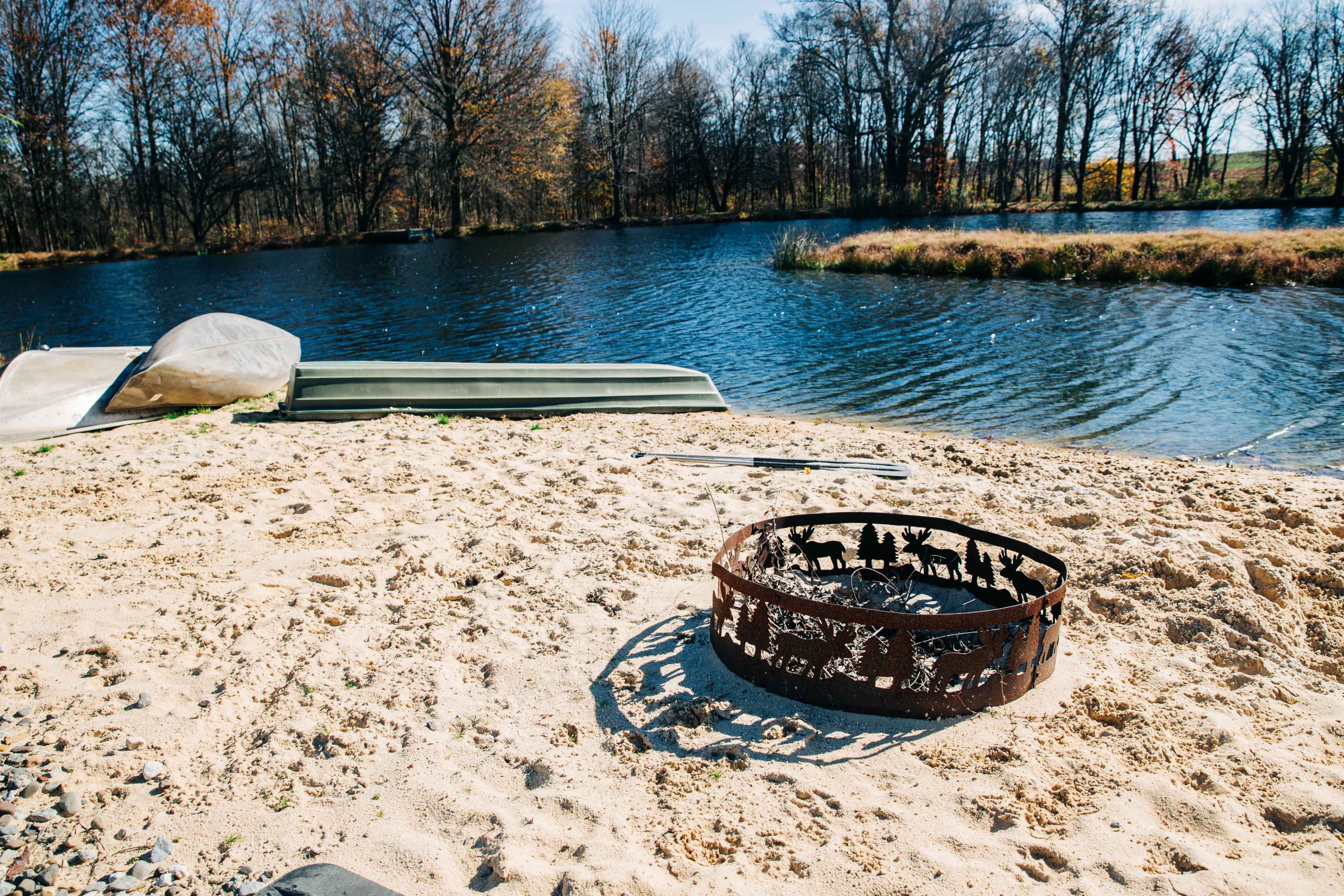 A fire pit made of metal is placed on a sandy area near a calm pond, with a canoe resting on the shore.
