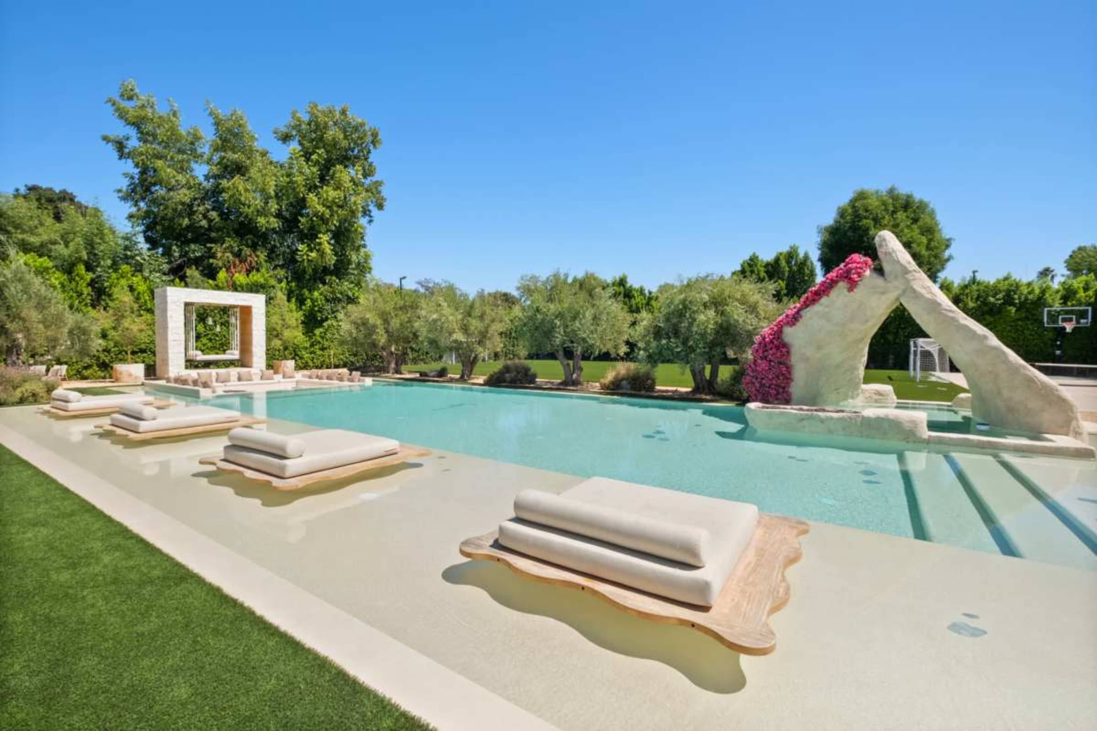 The image shows a modern swimming pool surrounded by lush greenery, featuring stone lounges and a decorative rock formation with plants.