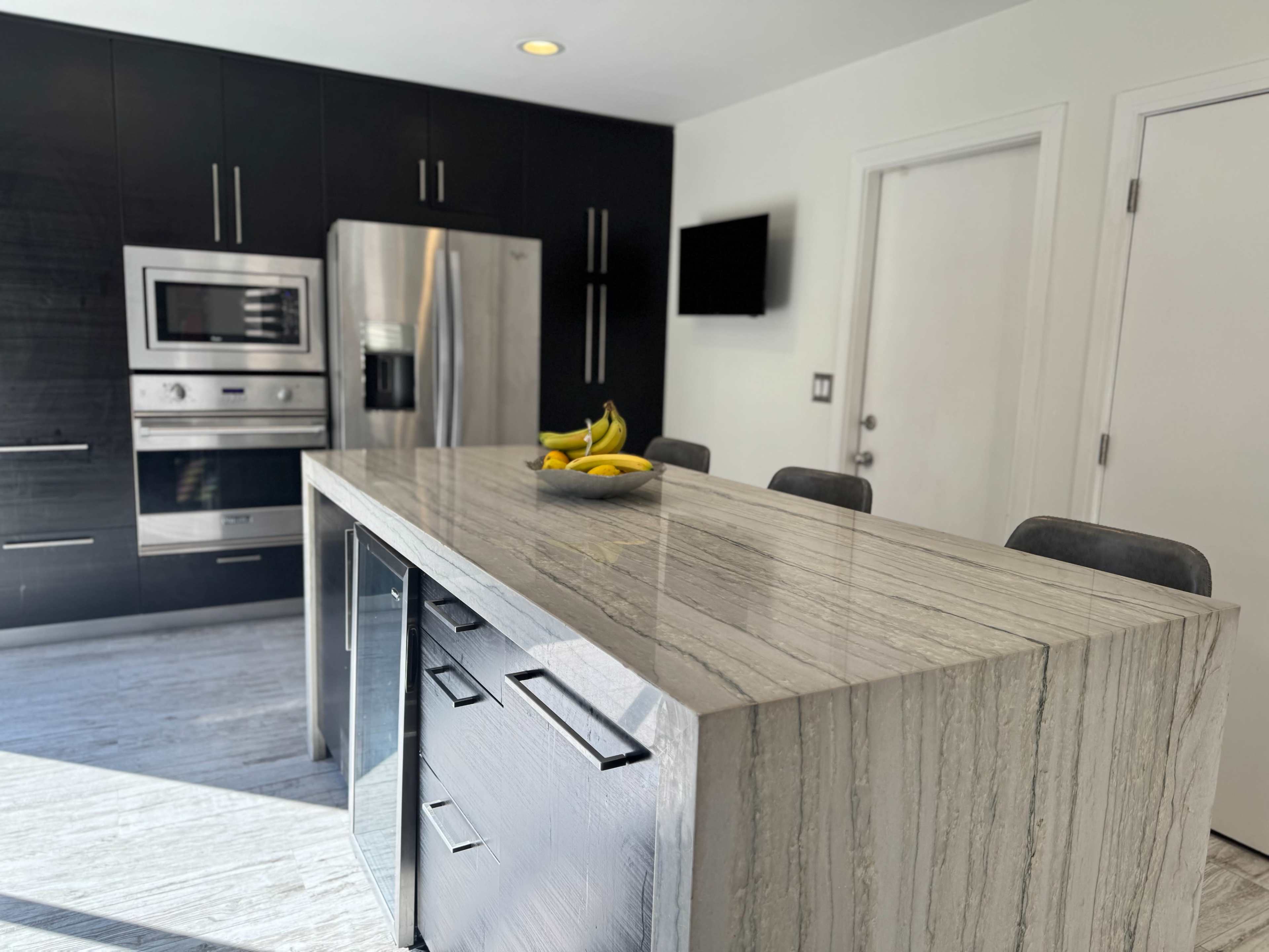 The image shows a modern kitchen featuring a large gray stone island with a bowl of bananas on top, stainless steel appliances, and dark cabinetry.