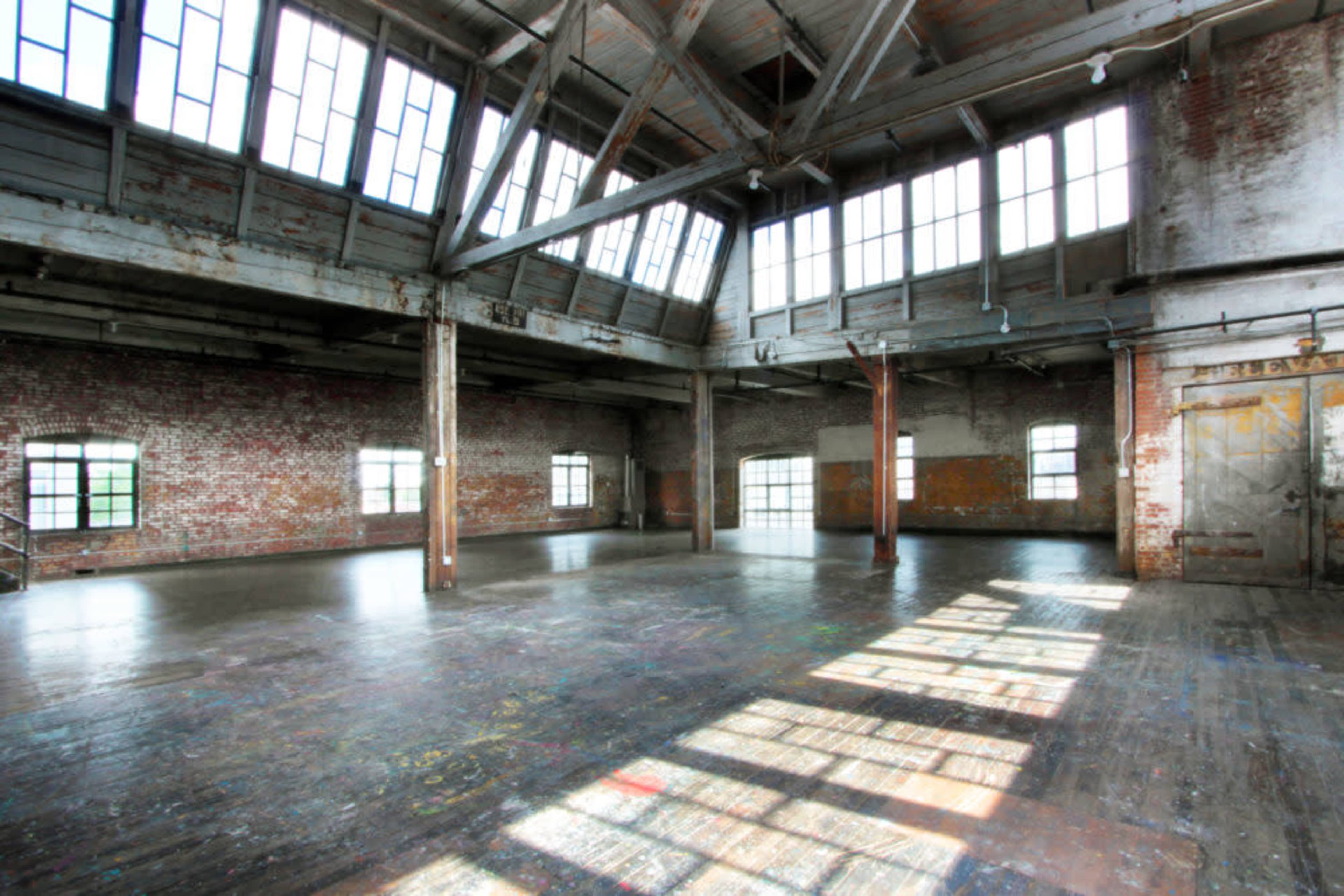 The image shows an empty industrial space with high ceilings, large windows, and exposed brick walls.