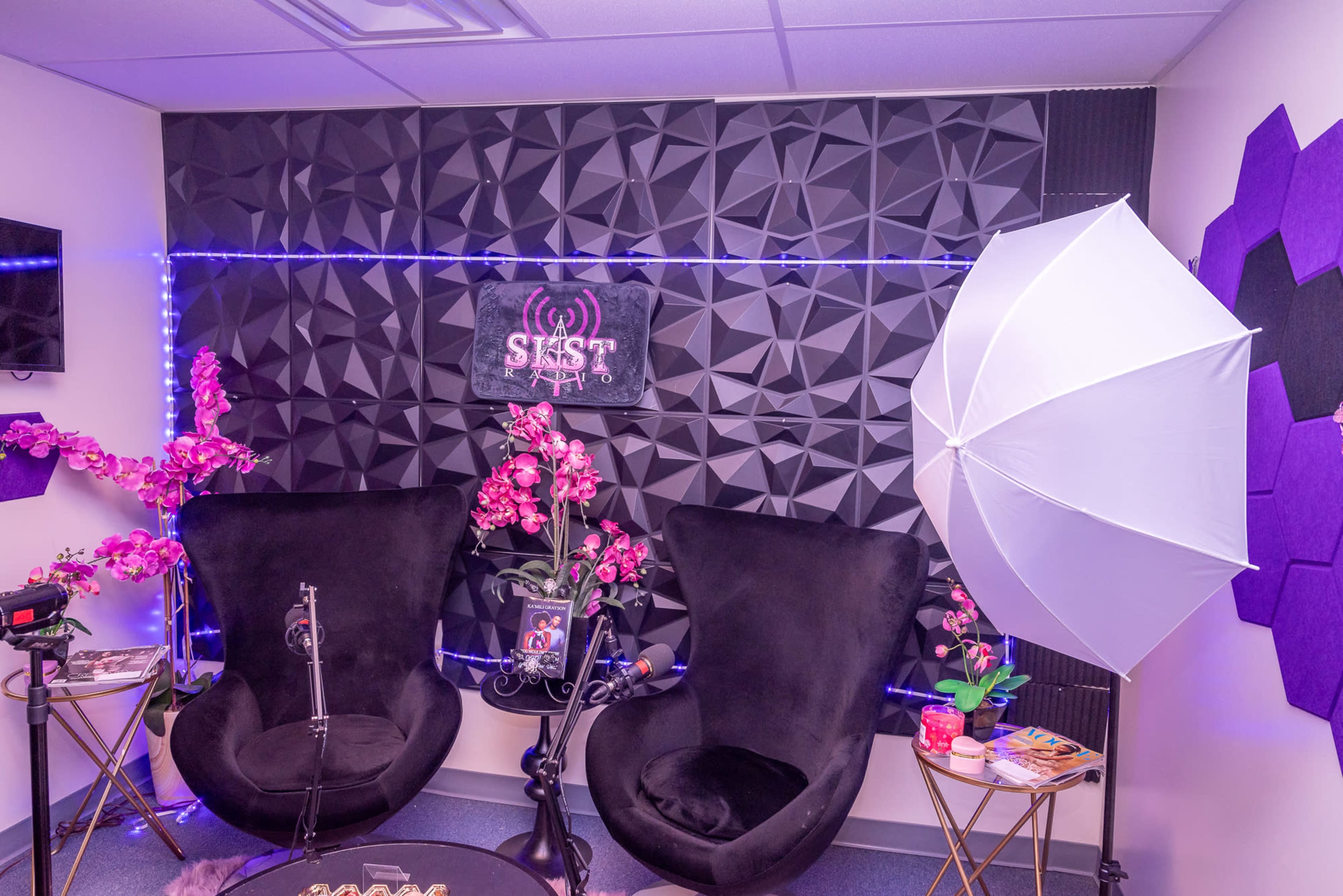 A podcast studio setup with two black armchairs, a microphone on a stand, decorative pink orchids, and an umbrella light against a textured wall.