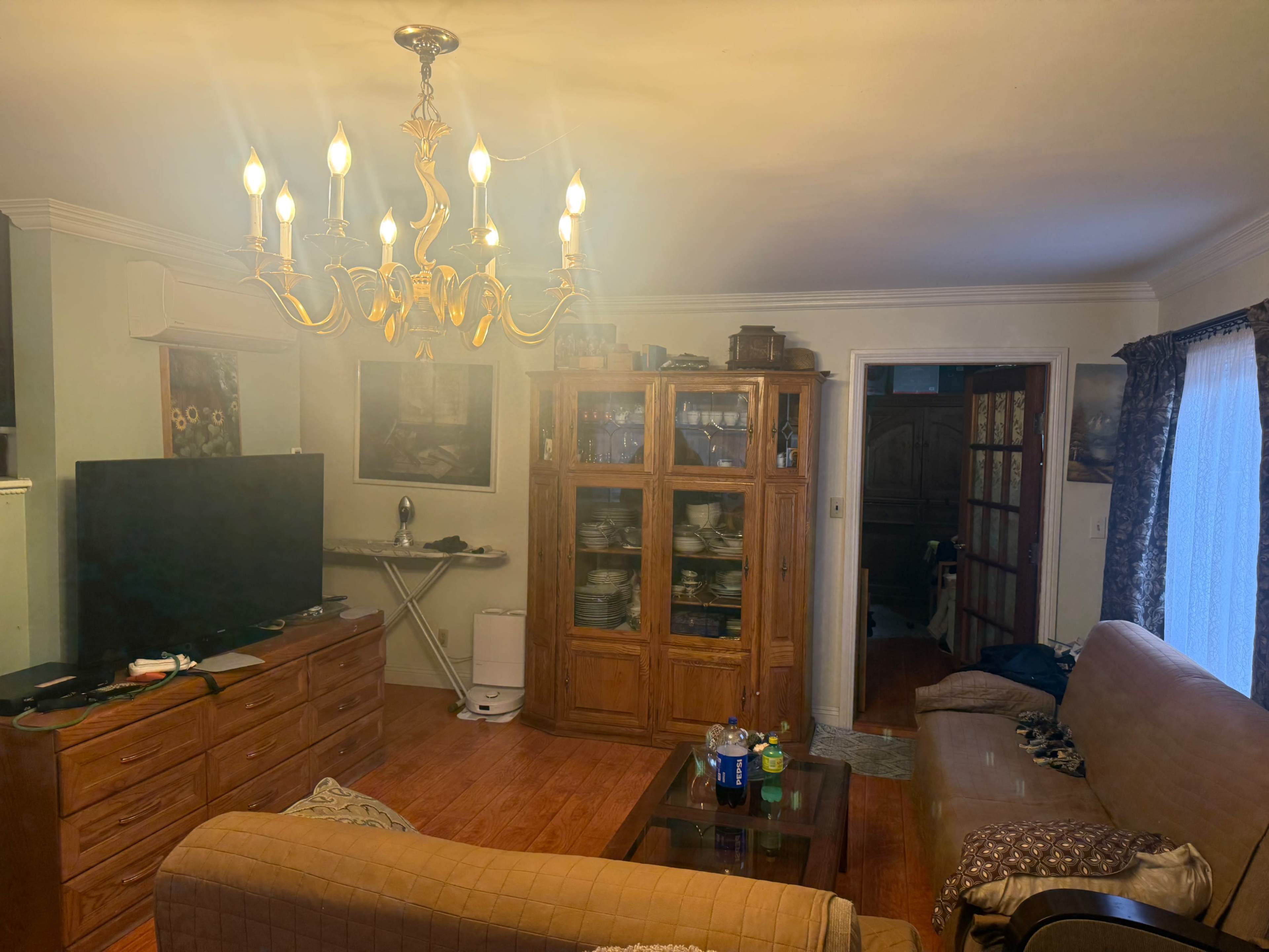 The living room features a chandelier, a wooden cabinet filled with glassware, a television on a wooden stand, and a doorway leading to another room.