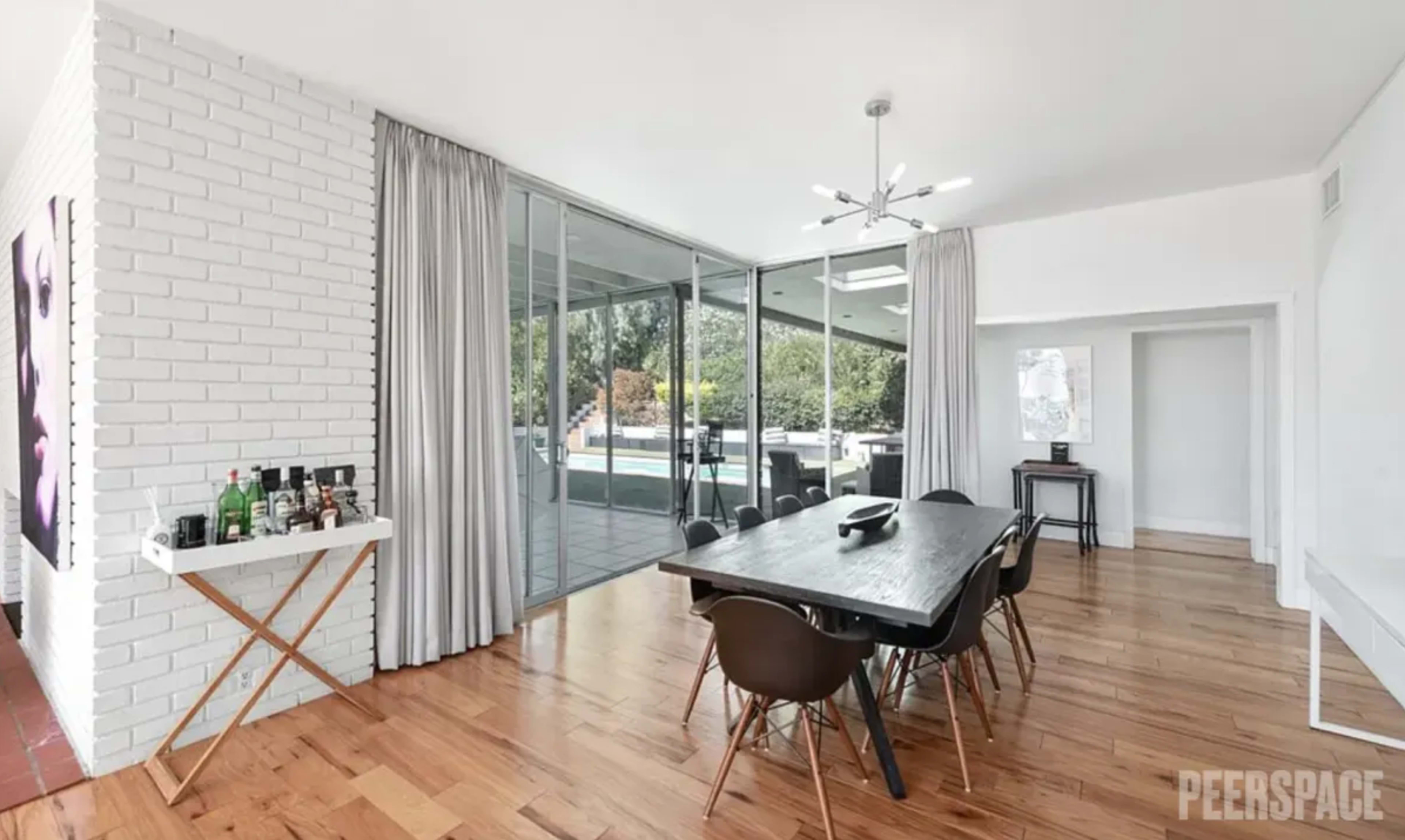 A modern dining area with a long table and chairs, large glass windows overlooking an outdoor pool, and white brick walls.