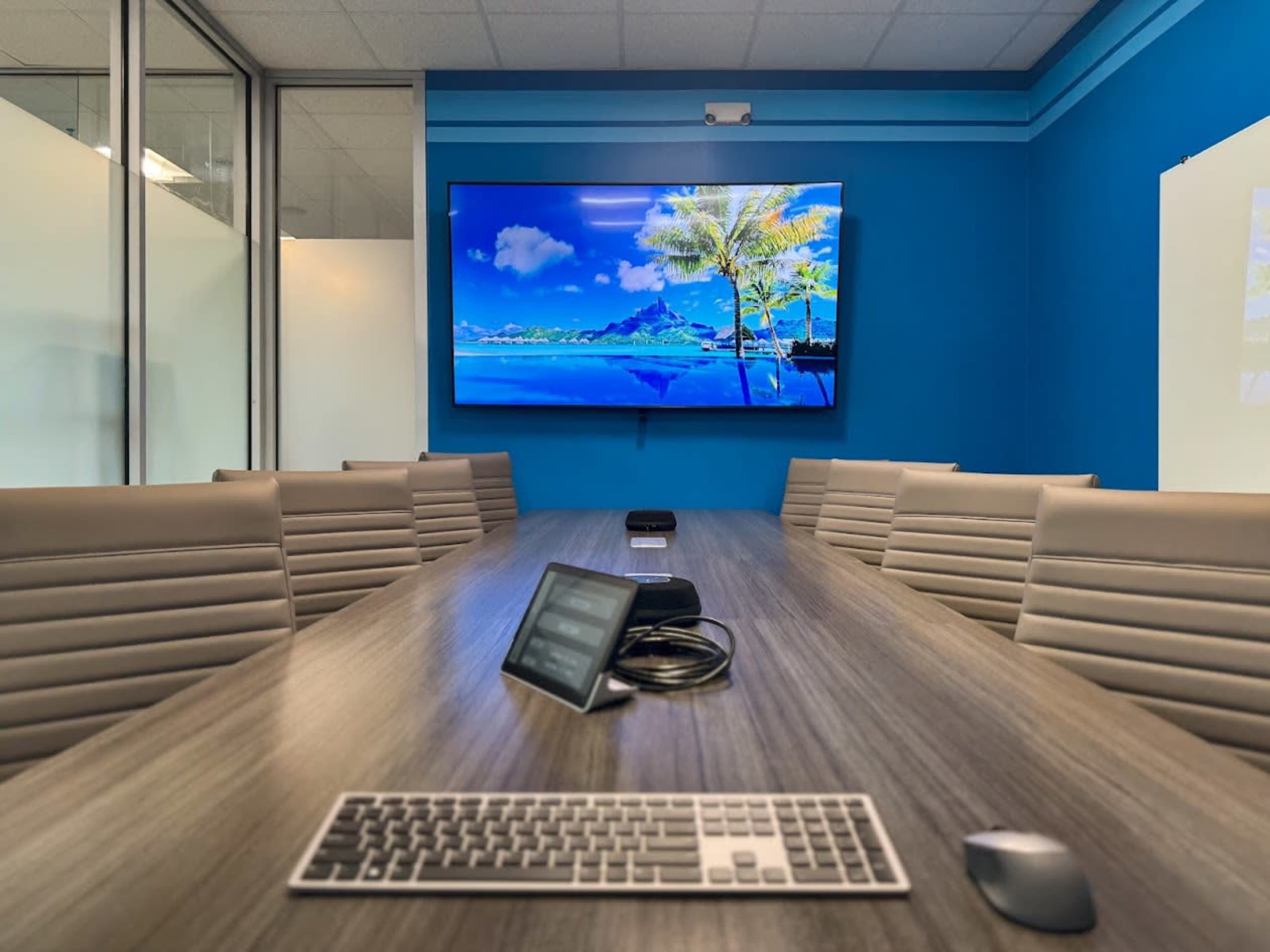 A modern conference room features a large table, a keyboard, and a screen displaying a tropical landscape on a blue-painted wall.