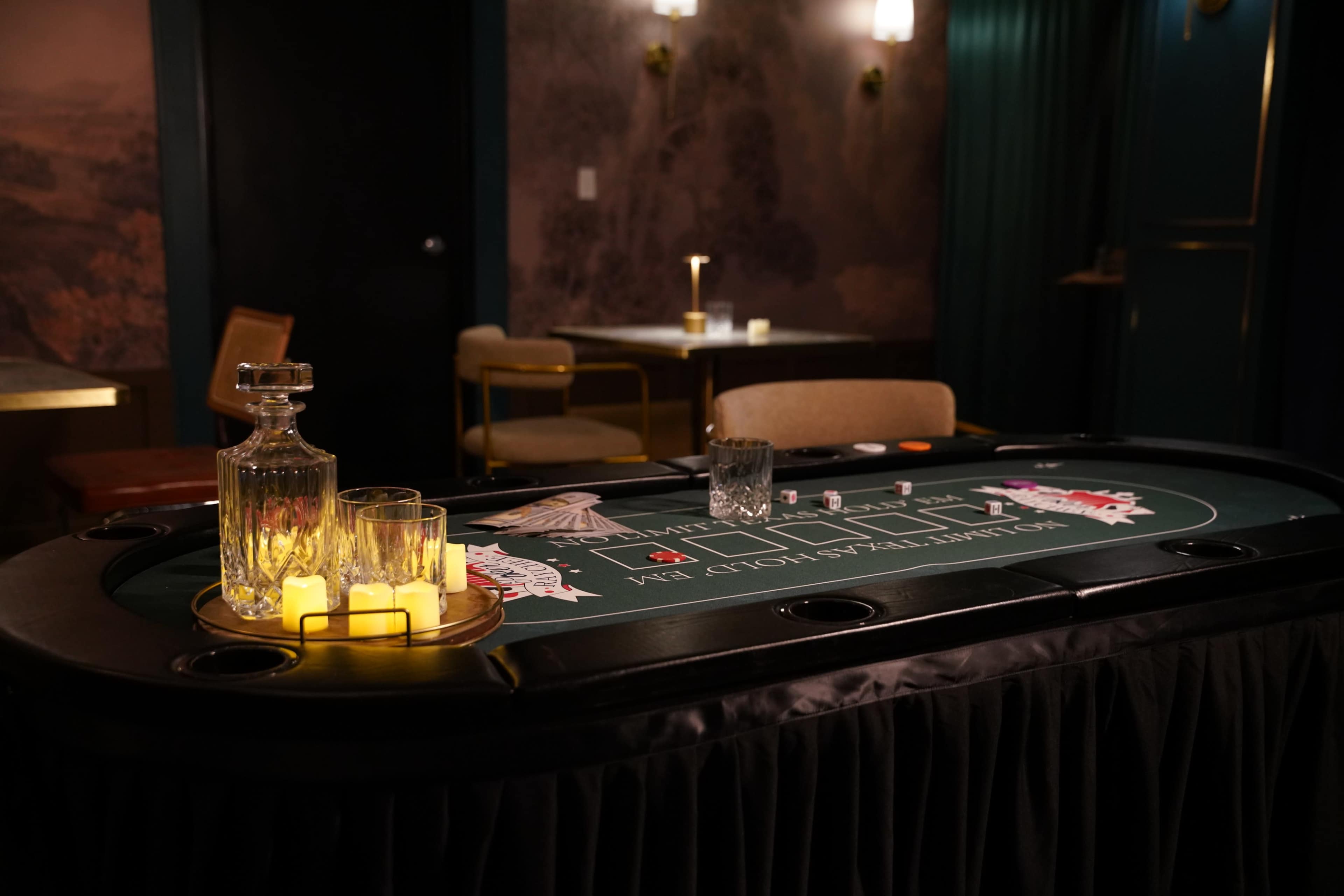 A casino table with a green felt layout, surrounded by chairs, features a decanter and glassware along with candles, set against a dimly lit backdrop.