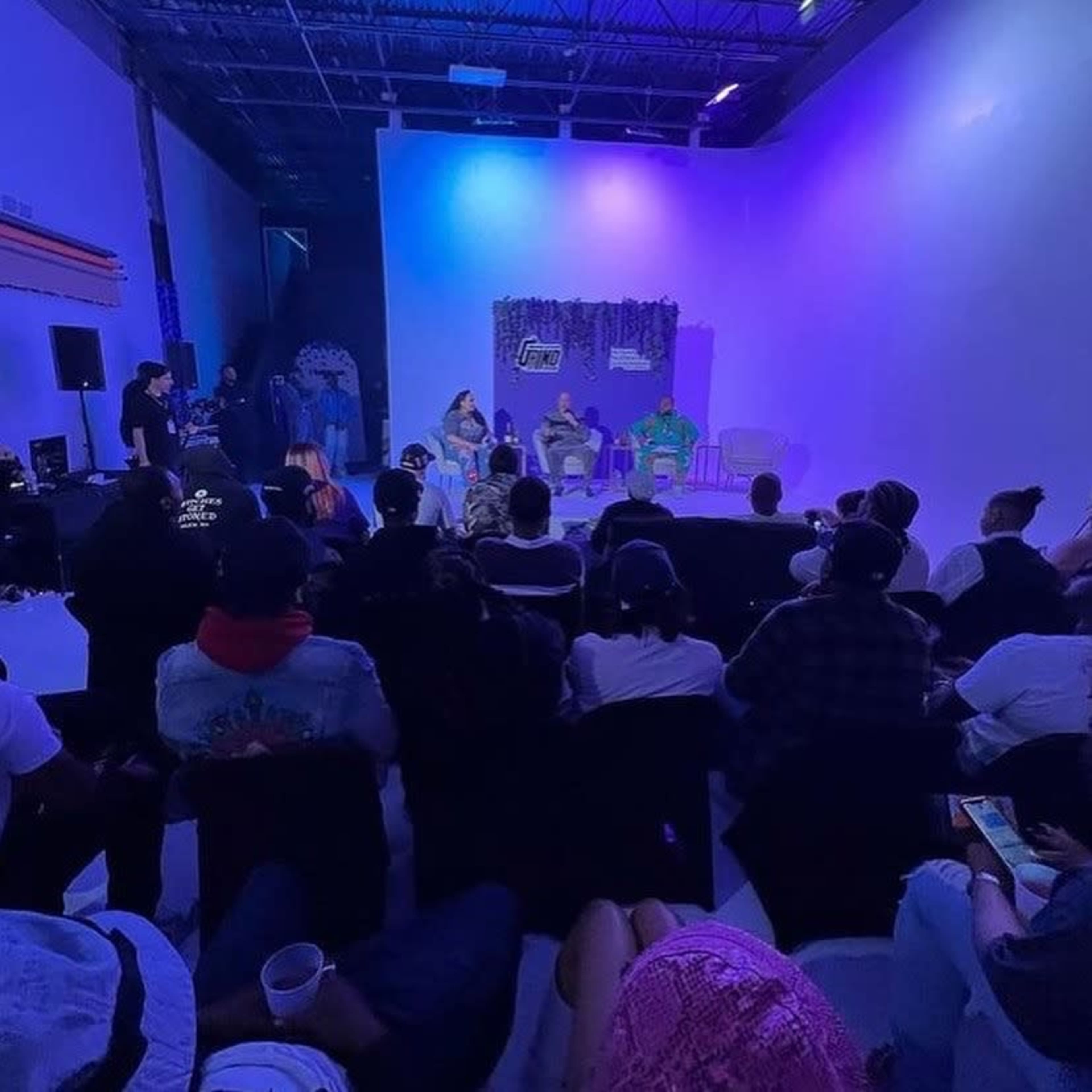 A group of people is seated in a dimly lit room, facing a small stage where three individuals are engaged in discussion.