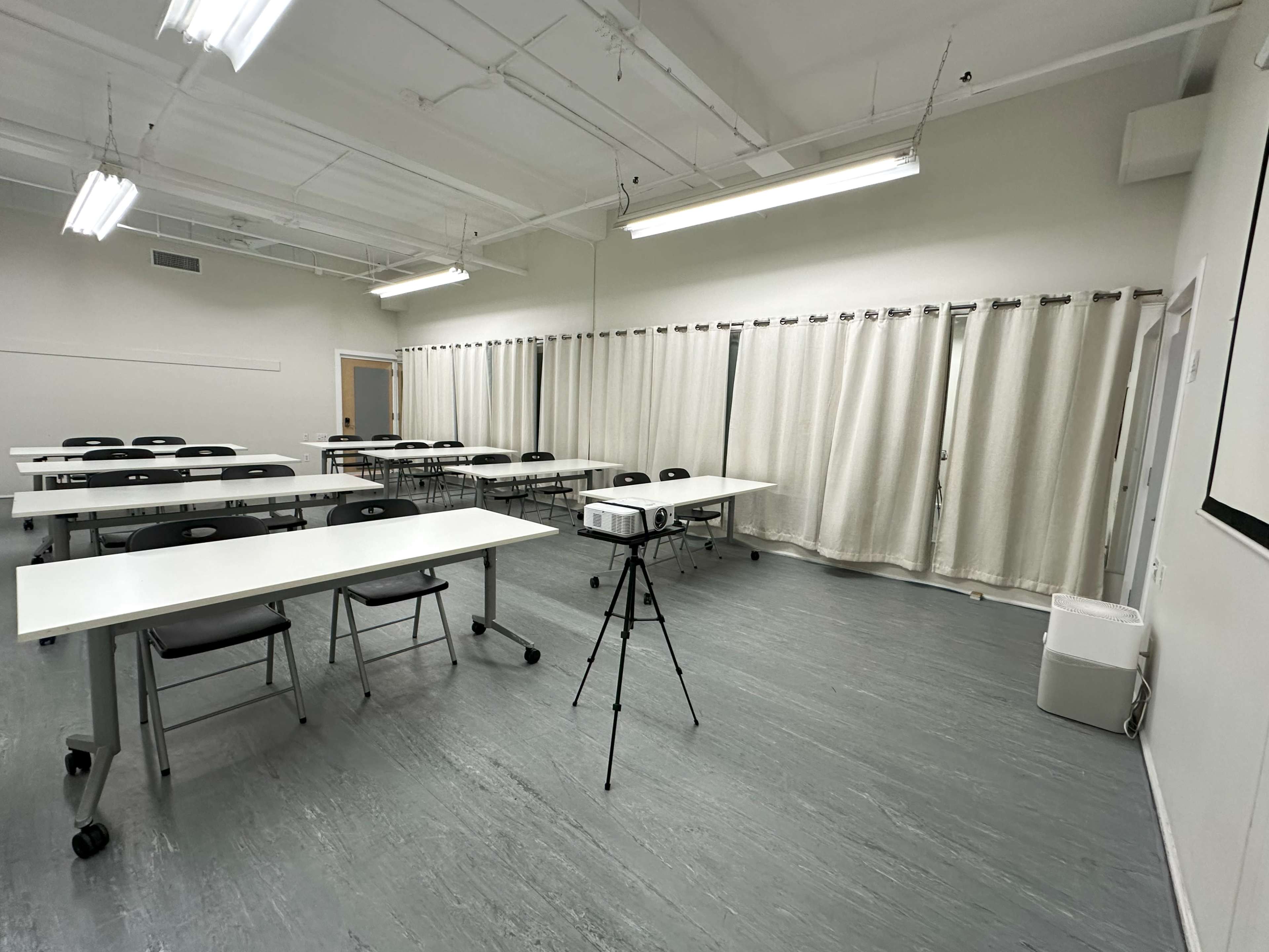 A spacious, empty classroom with several tables arranged in rows and a projector on a tripod in the center.