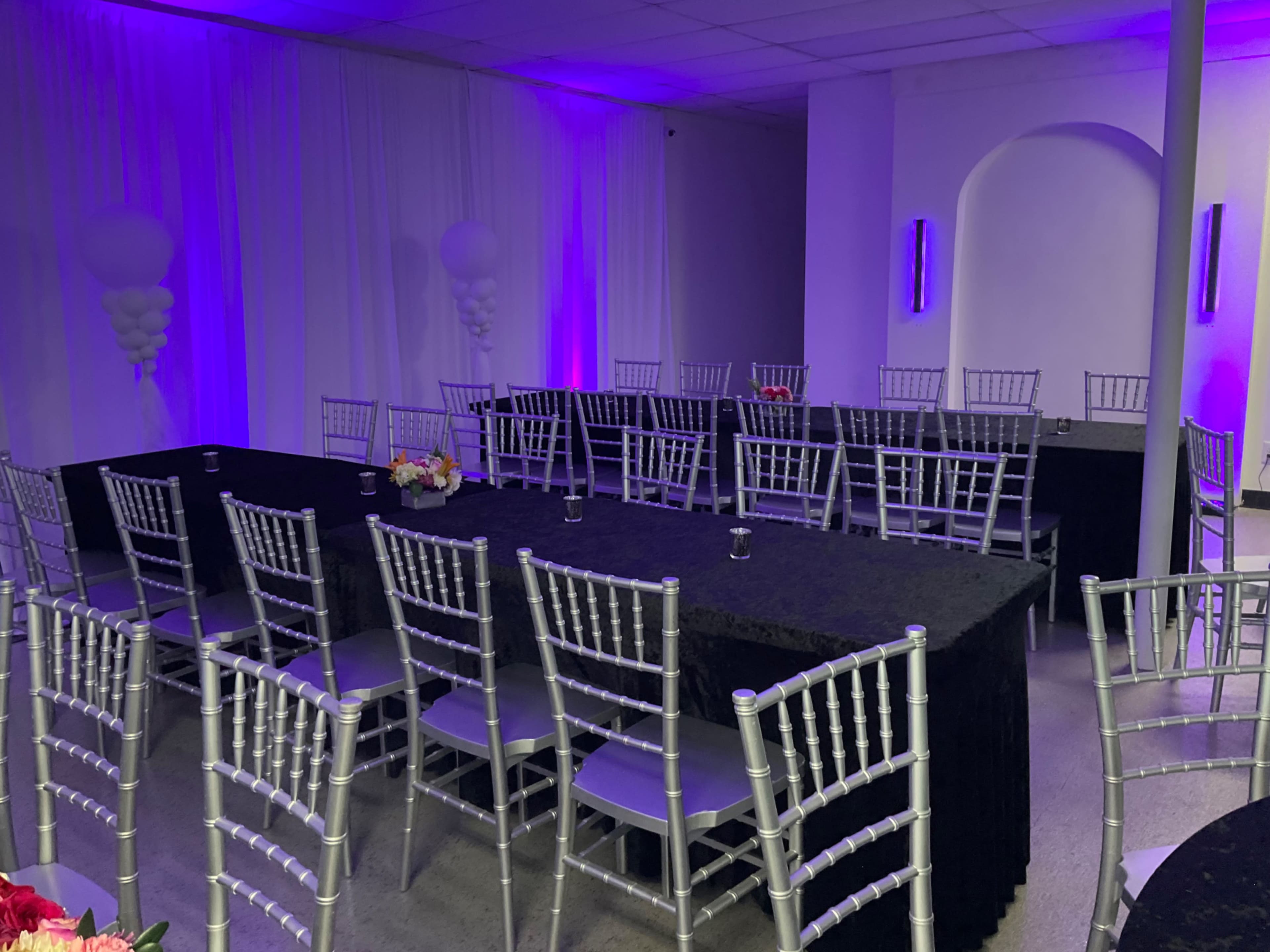 A banquet hall is set with tables covered in black cloths and silver chiavari chairs, illuminated by purple lighting.