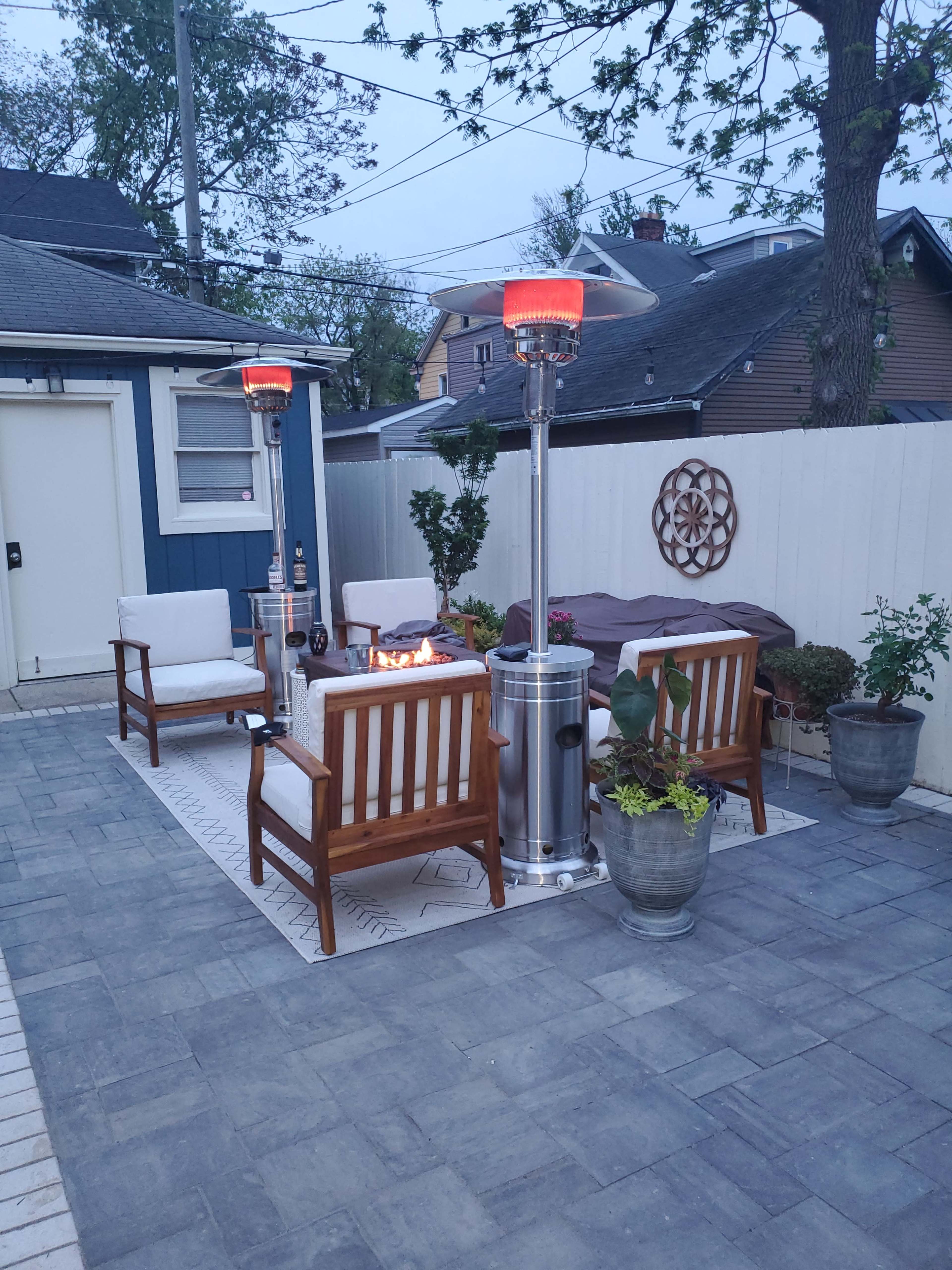 A cozy outdoor seating area features two patio heaters, four wooden chairs, a fire pit, and potted plants on a stone patio.