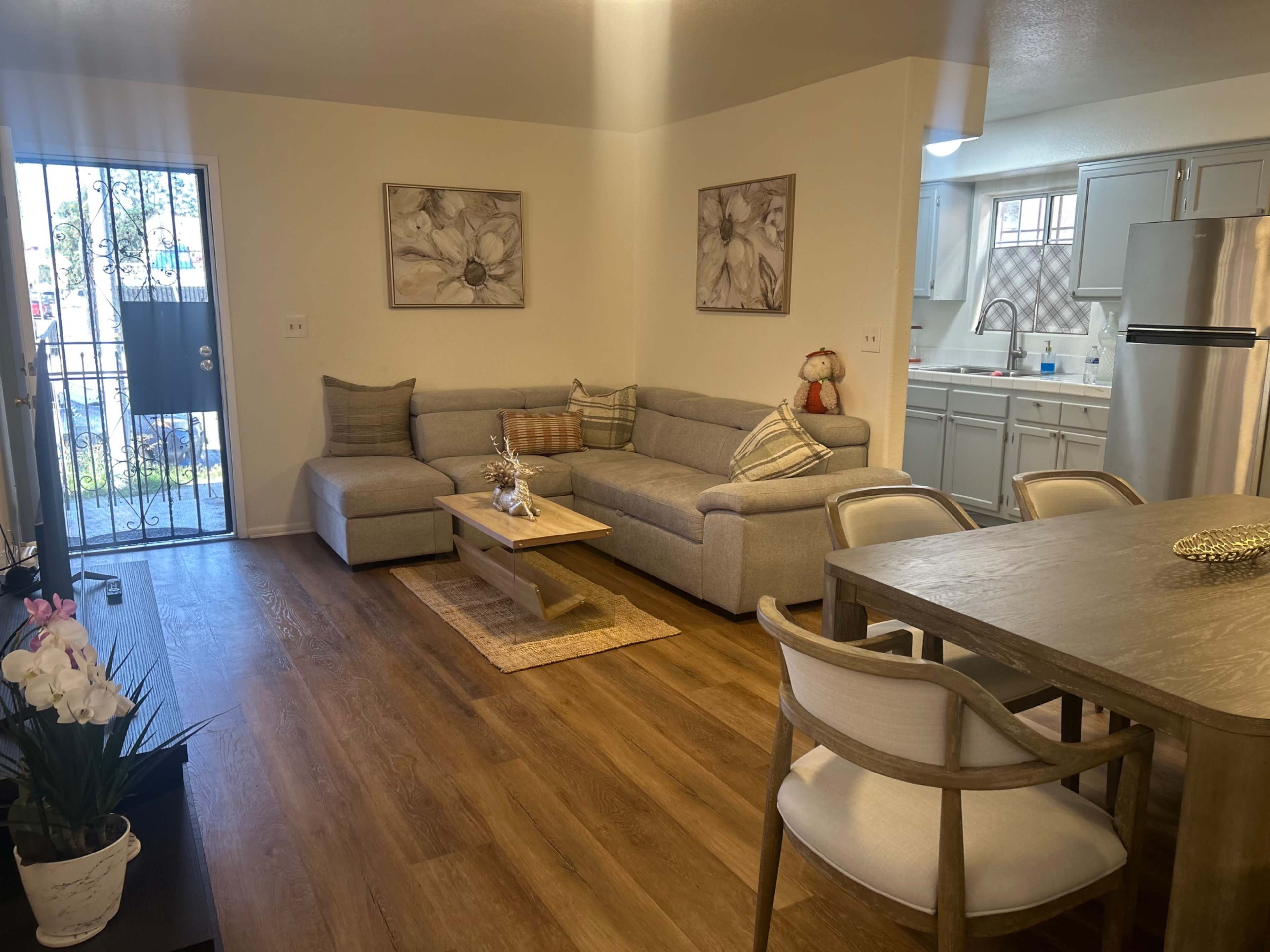 The image shows a modern living area featuring a gray sectional couch, a wooden coffee table, and a dining space with a light wood table and chairs, leading to a kitchen area in the background.
