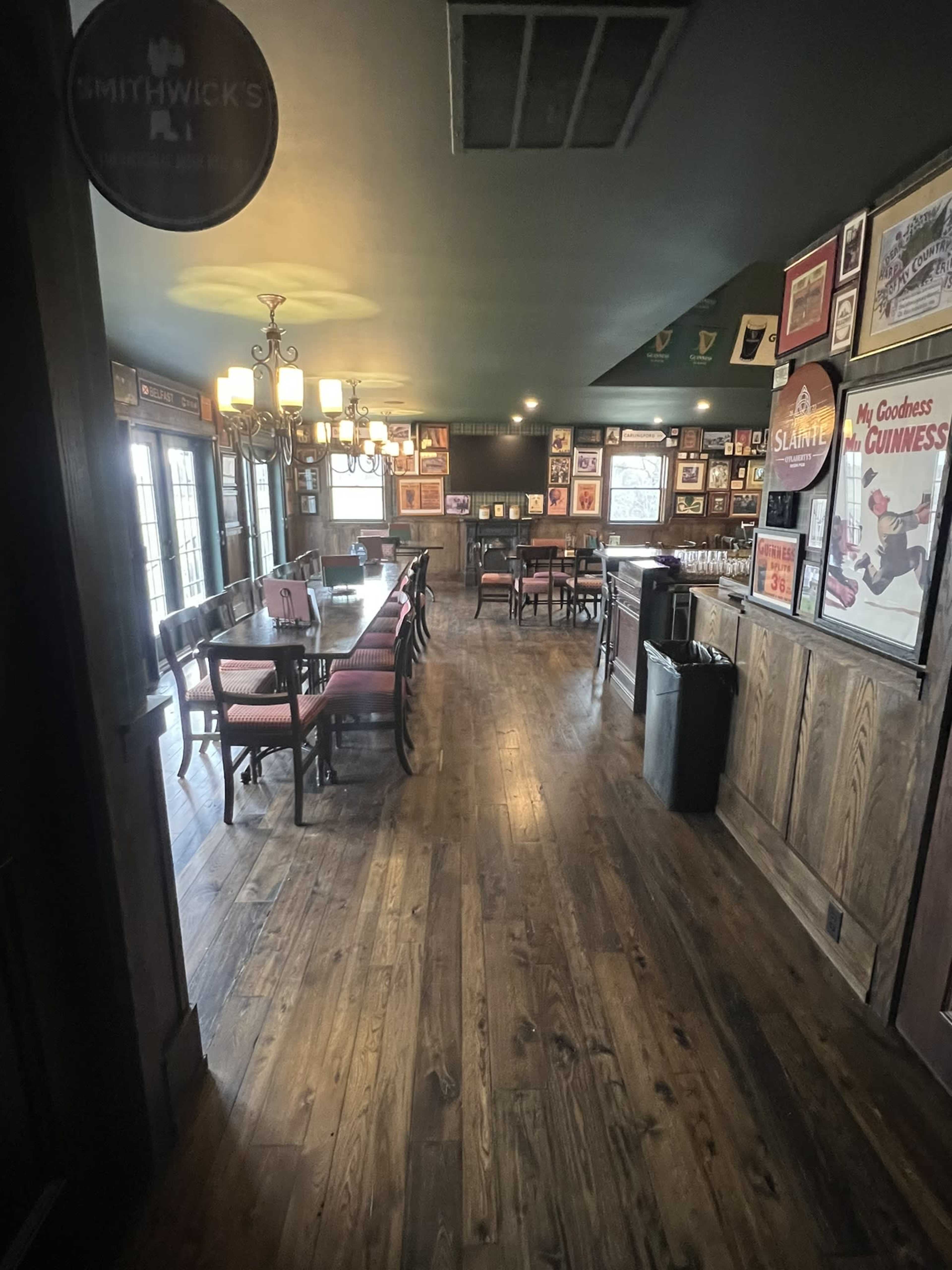The image shows a dimly lit pub interior with wooden flooring, a long table set for dining, and various framed posters and artworks on the walls.