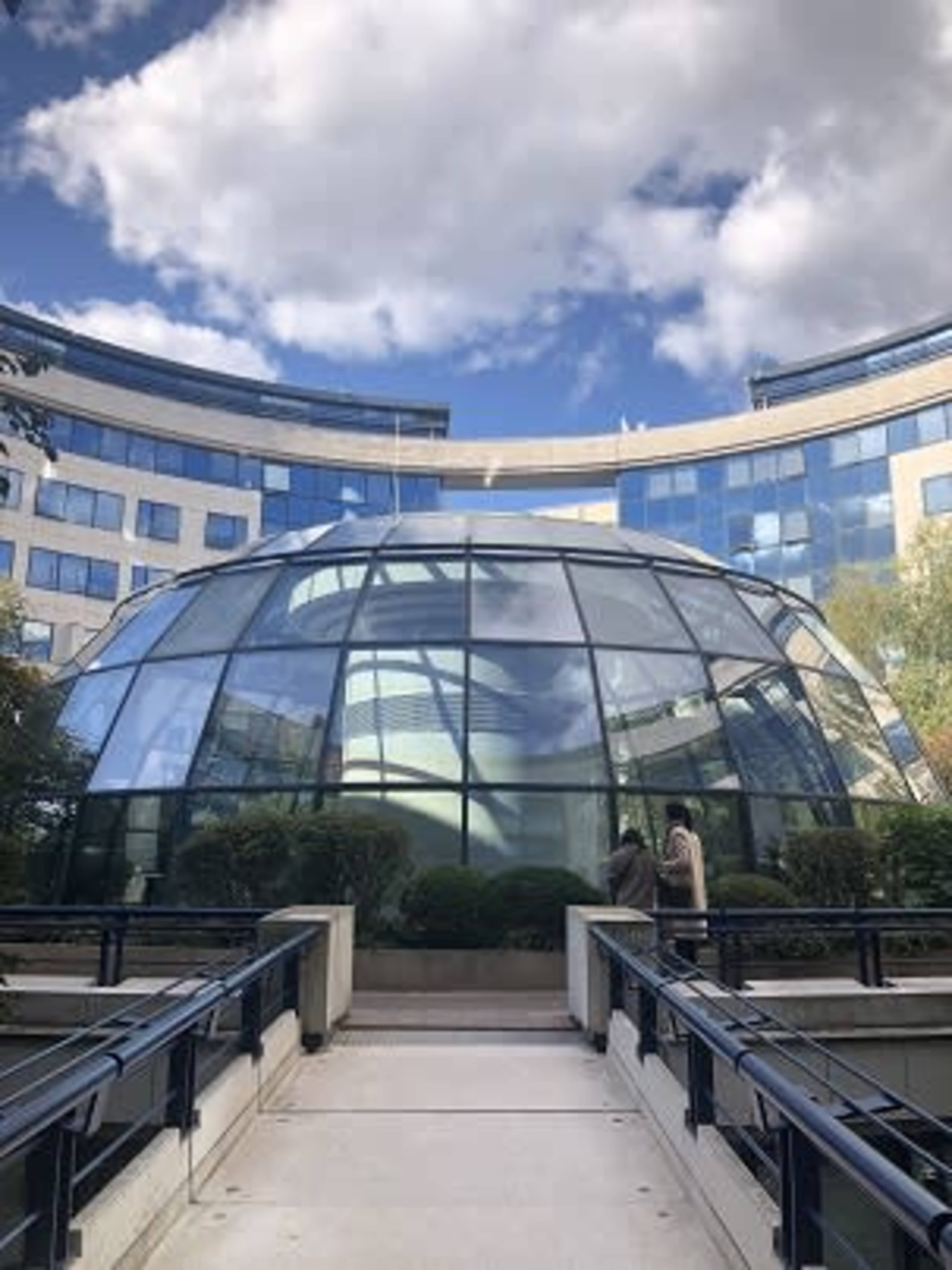 A glass dome structure is situated at the entrance of a modern building surrounded by greenery and a pathway.