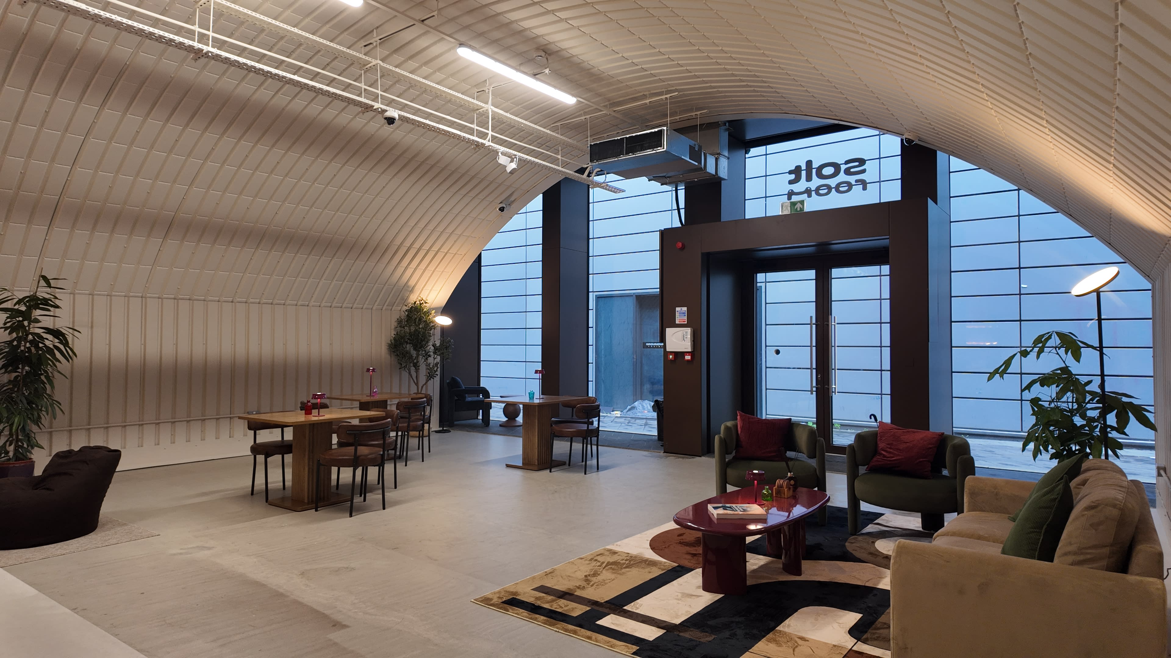 The image shows a modern, spacious lounge area with tables, chairs, and plants, featuring large windows and a distinctive arched ceiling.