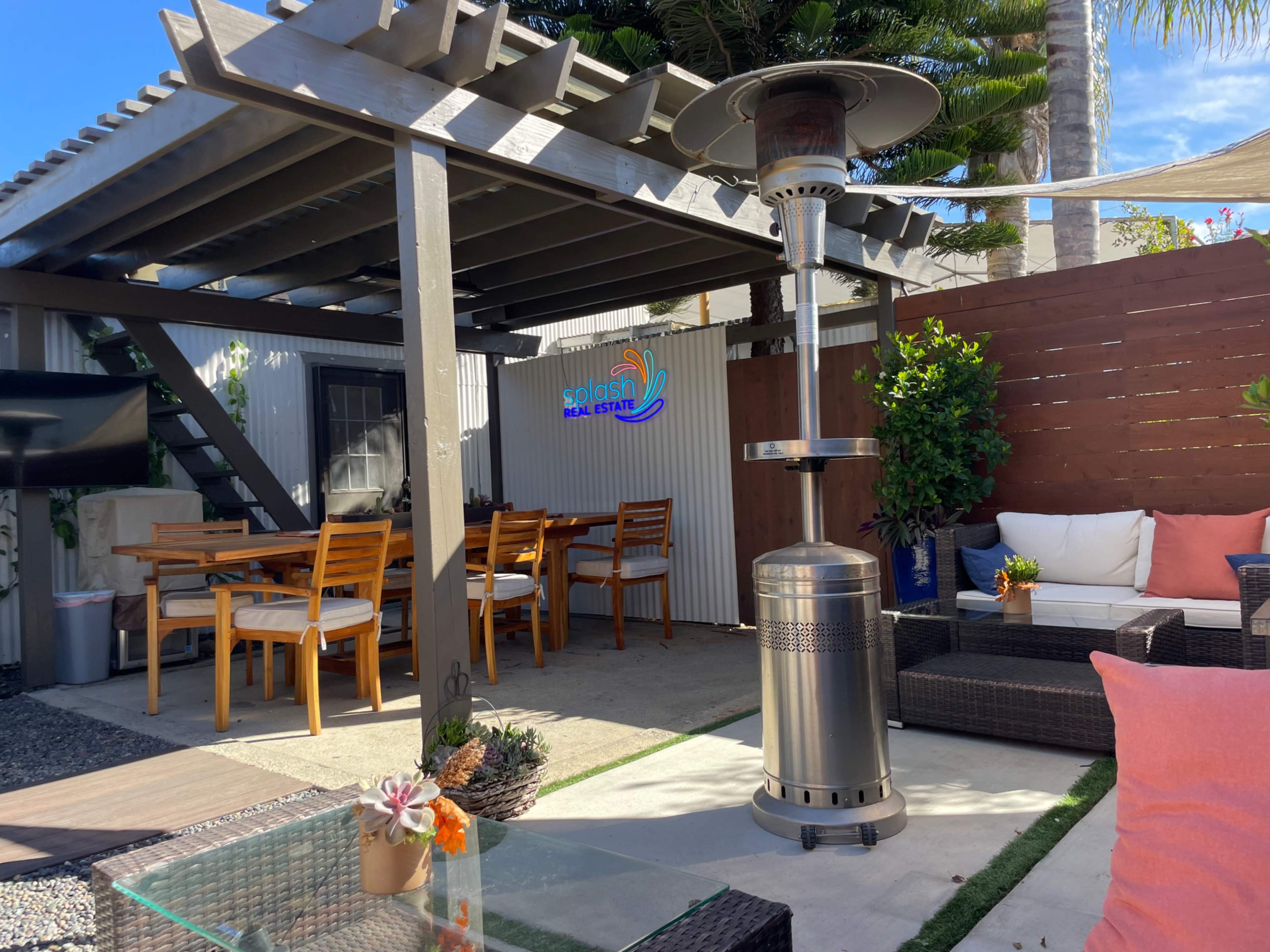 A patio area features a dining table, comfortable seating, and a patio heater under a pergola surrounded by greenery.