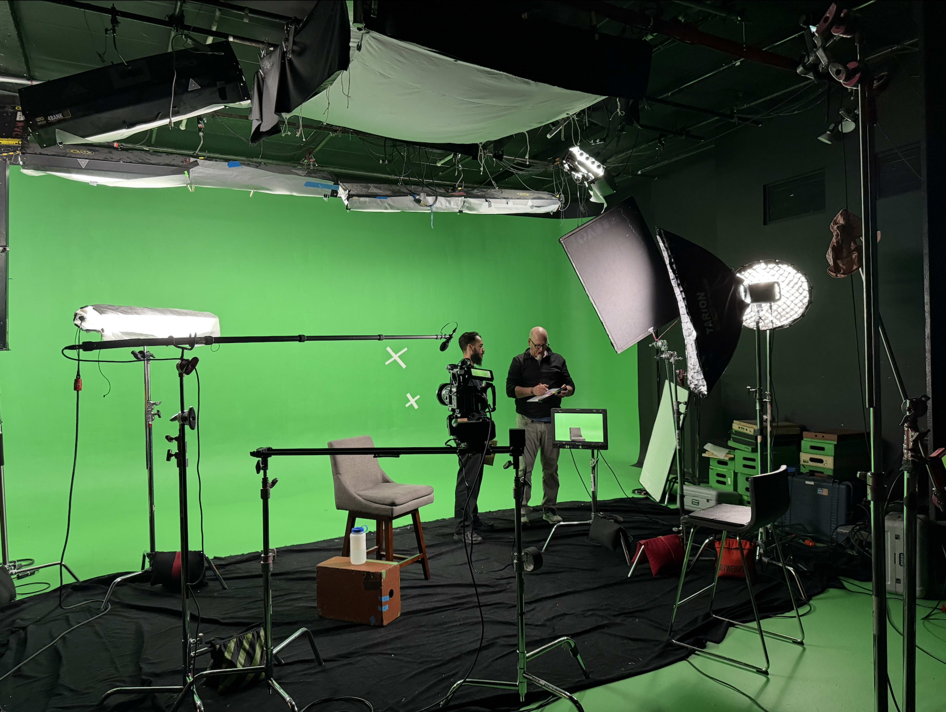 A green screen studio setup features two men preparing equipment, with various lighting and camera gear arranged around empty chairs and a backdrop.
