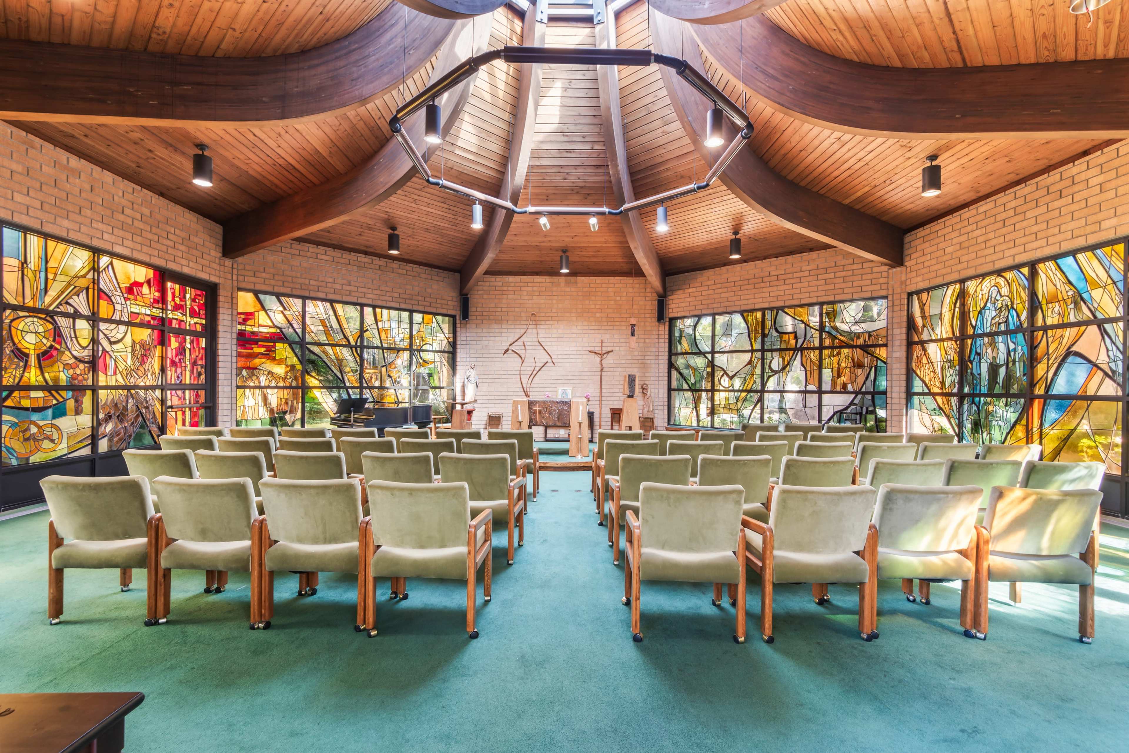 The image shows the interior of a chapel with rows of green chairs, a wooden ceiling, and large stained glass windows.