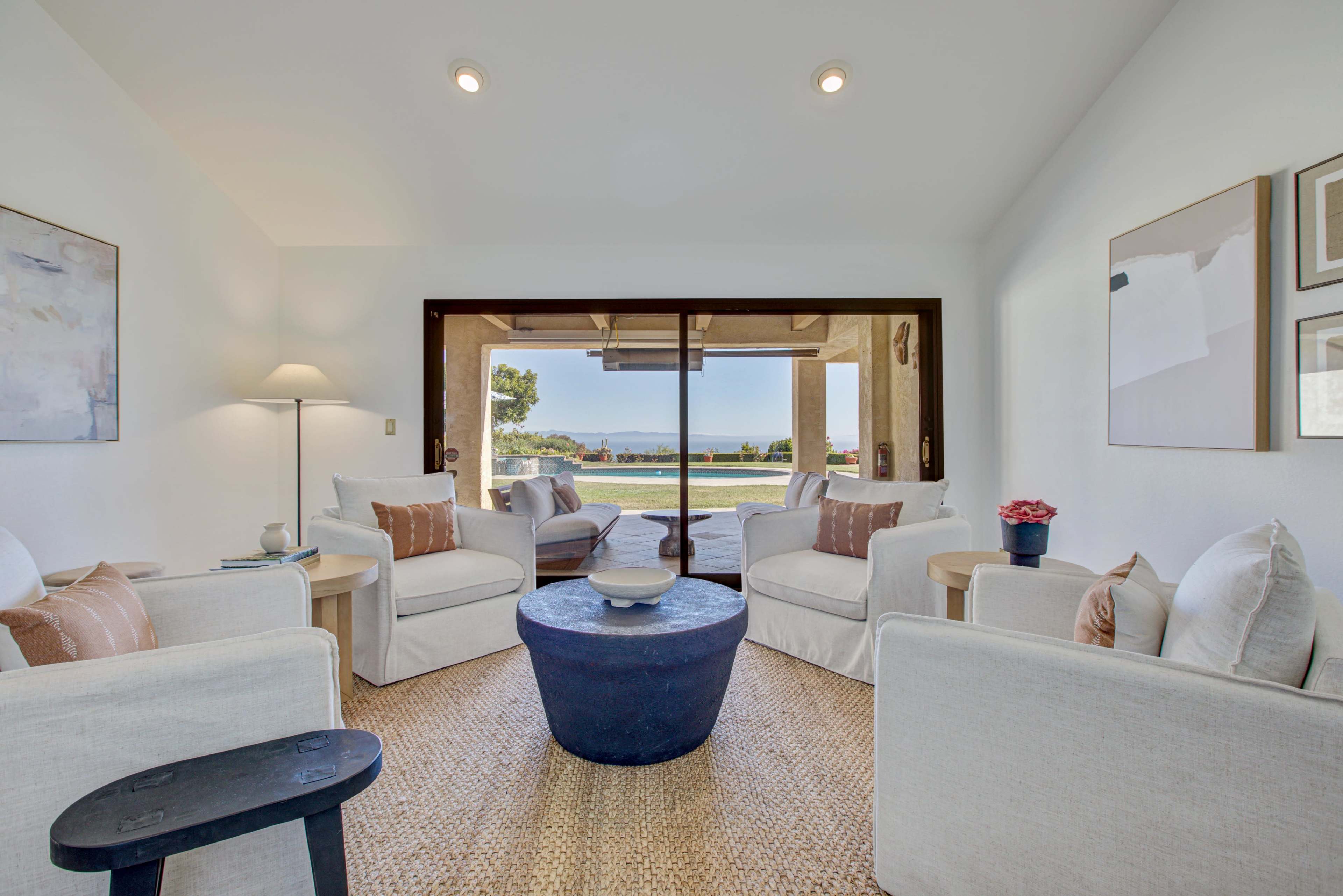 A bright living room features two white chairs and a blue coffee table, with large glass doors opening to a view of a landscaped outdoor area.
