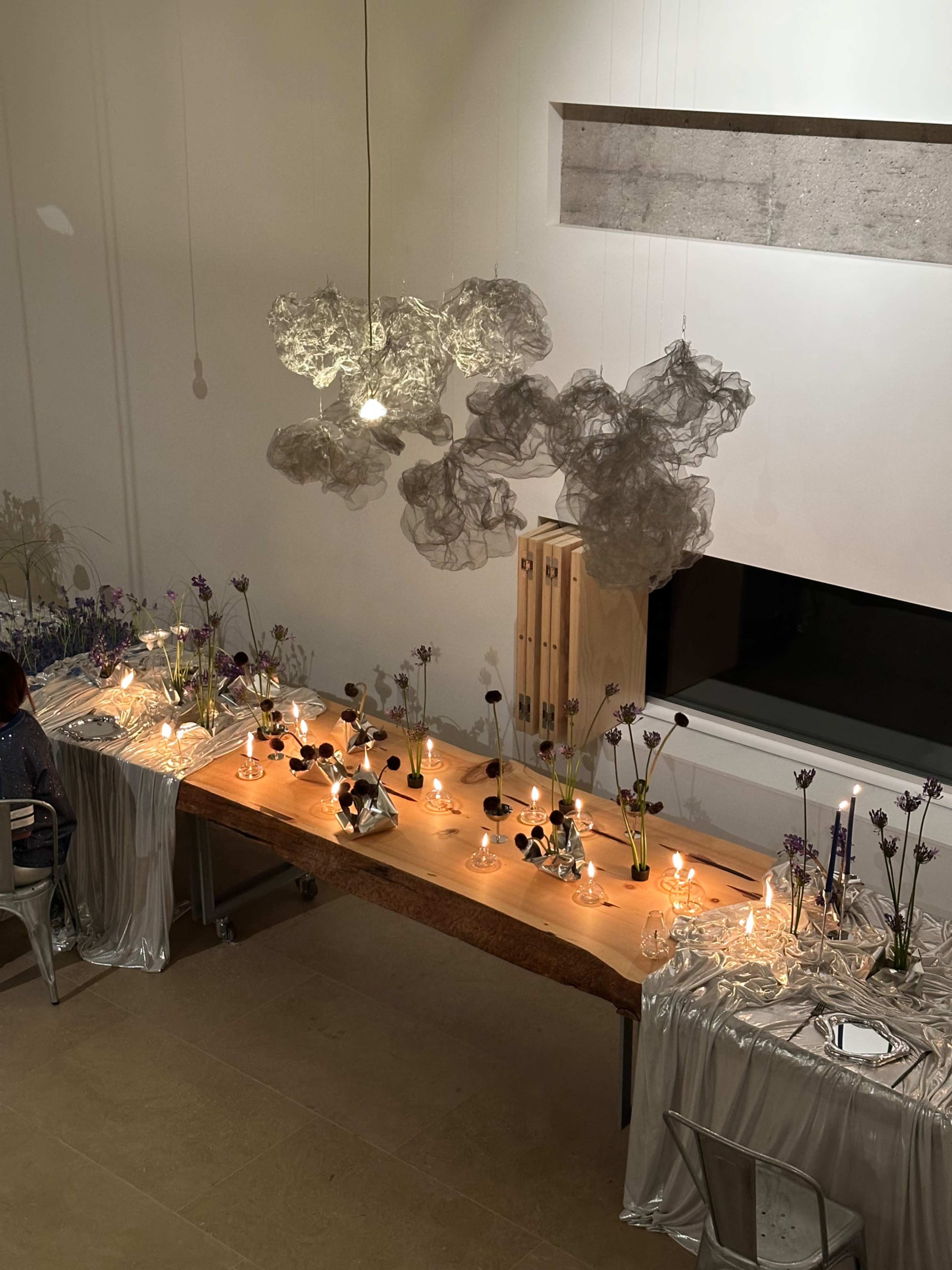 A long, elegantly set dining table with candles and flowers is illuminated by soft lighting, and decorative cloud-like sculptures hang above.