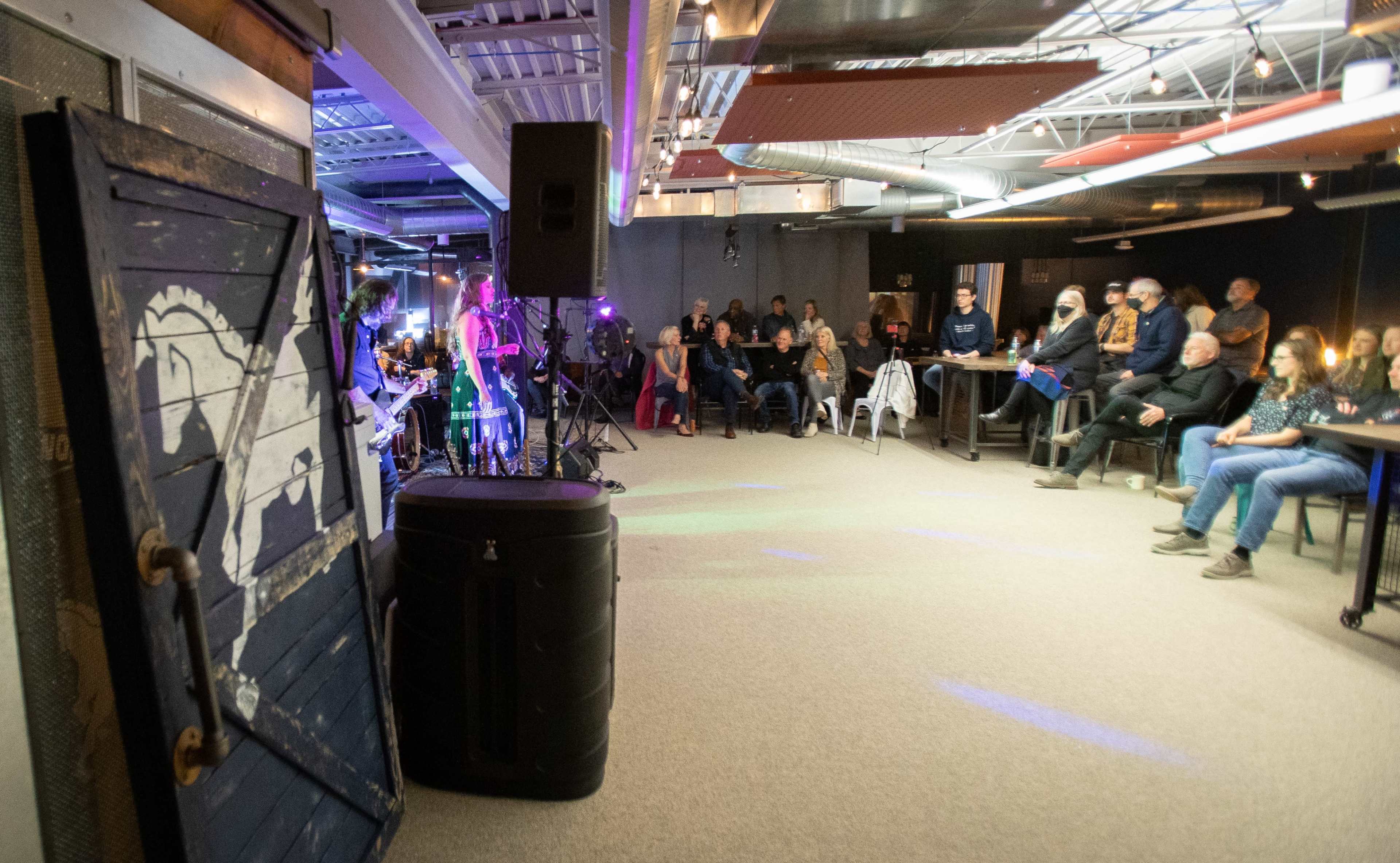 A musician performs on stage in a dimly lit venue while an audience sits attentively at tables.