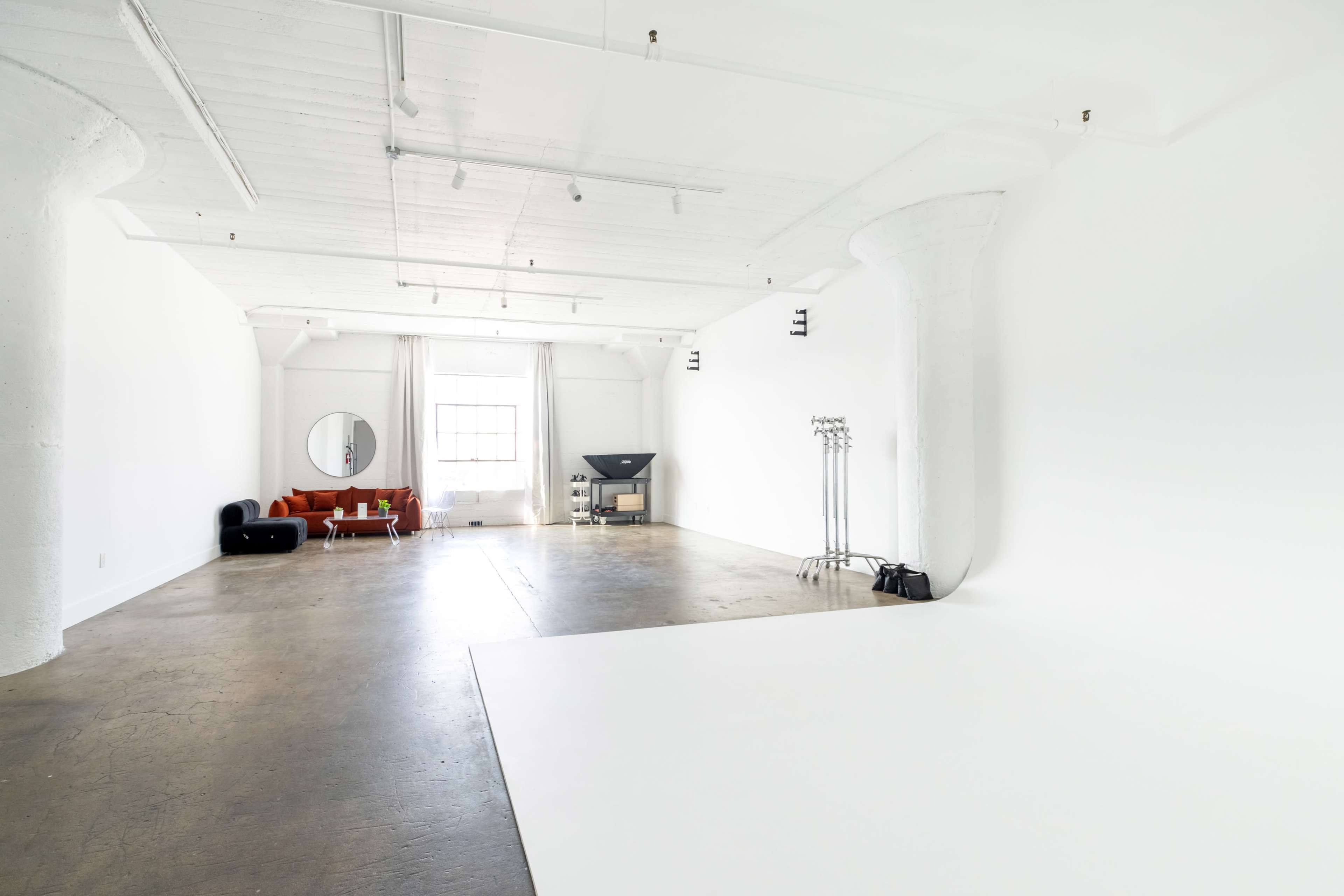 The image shows a spacious, empty studio with white walls, a concrete floor, a round mirror, and minimal furniture including a red couch and a television.