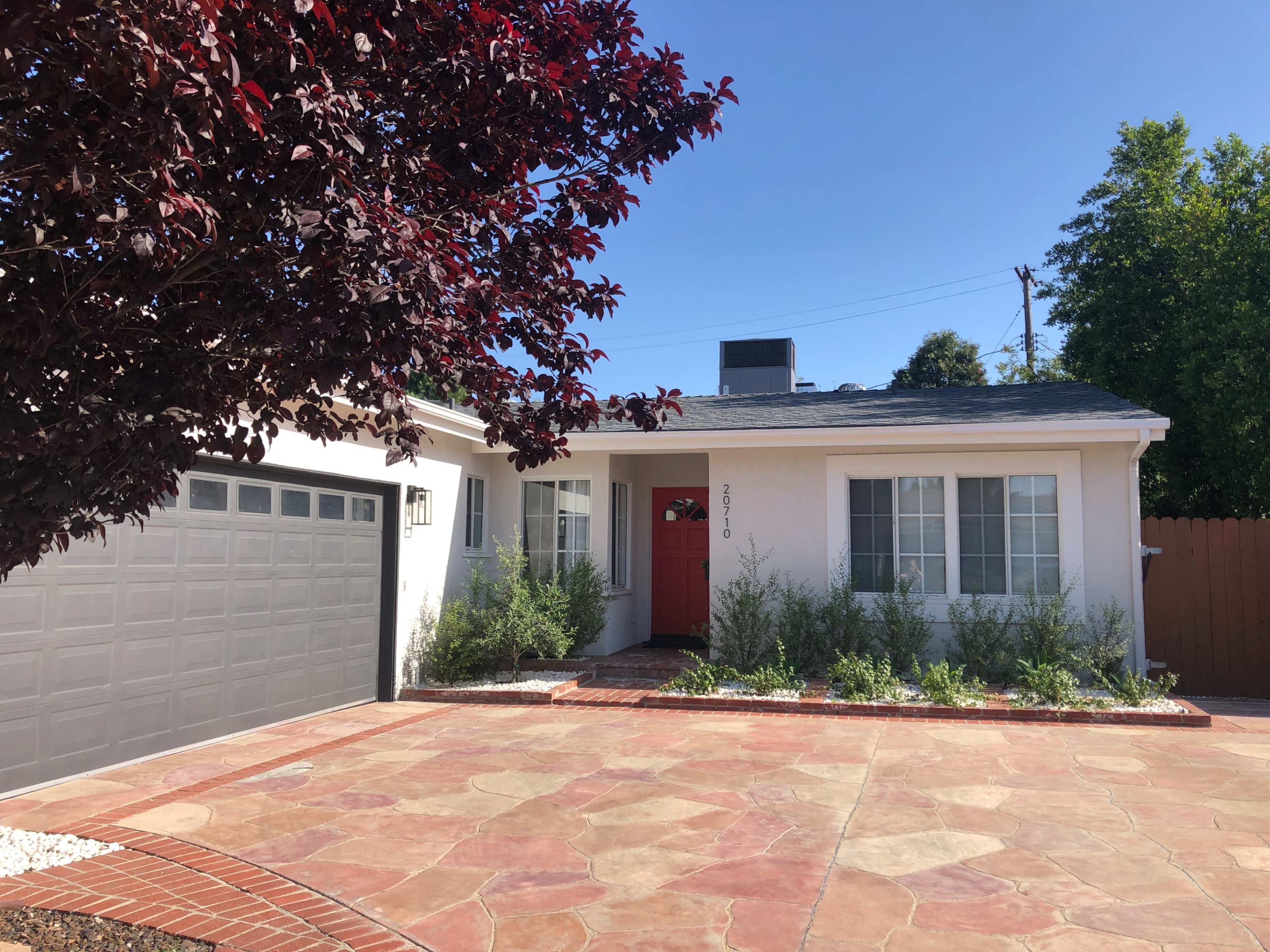 A single-story white house with a red front door and a gray garage sits on a stone driveway, bordered by greenery and a decorative tree.