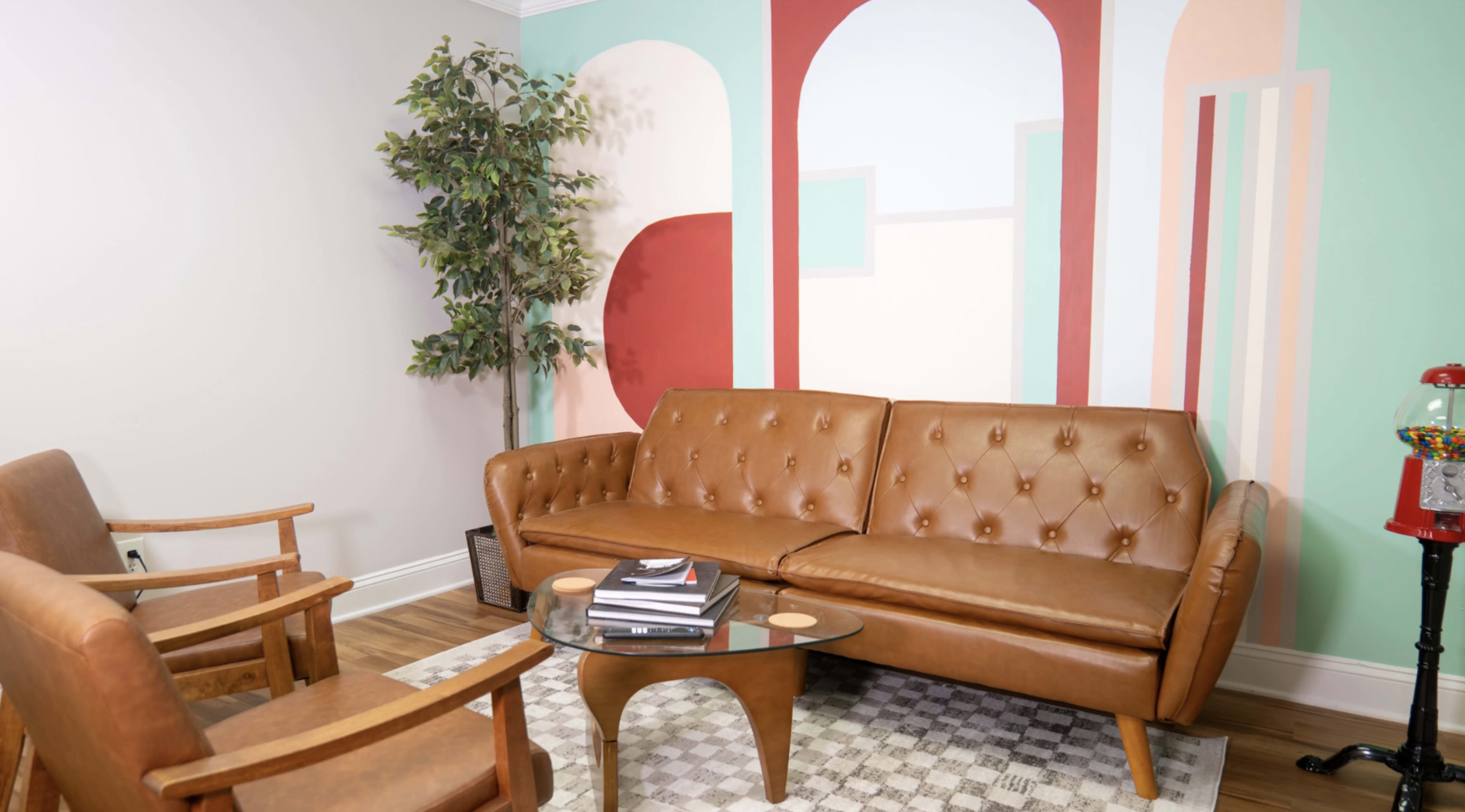 The image shows a cozy interior space featuring a brown tufted leather sofa, a glass coffee table with books, and a plant in a corner, alongside a brightly colored abstract mural on the wall.