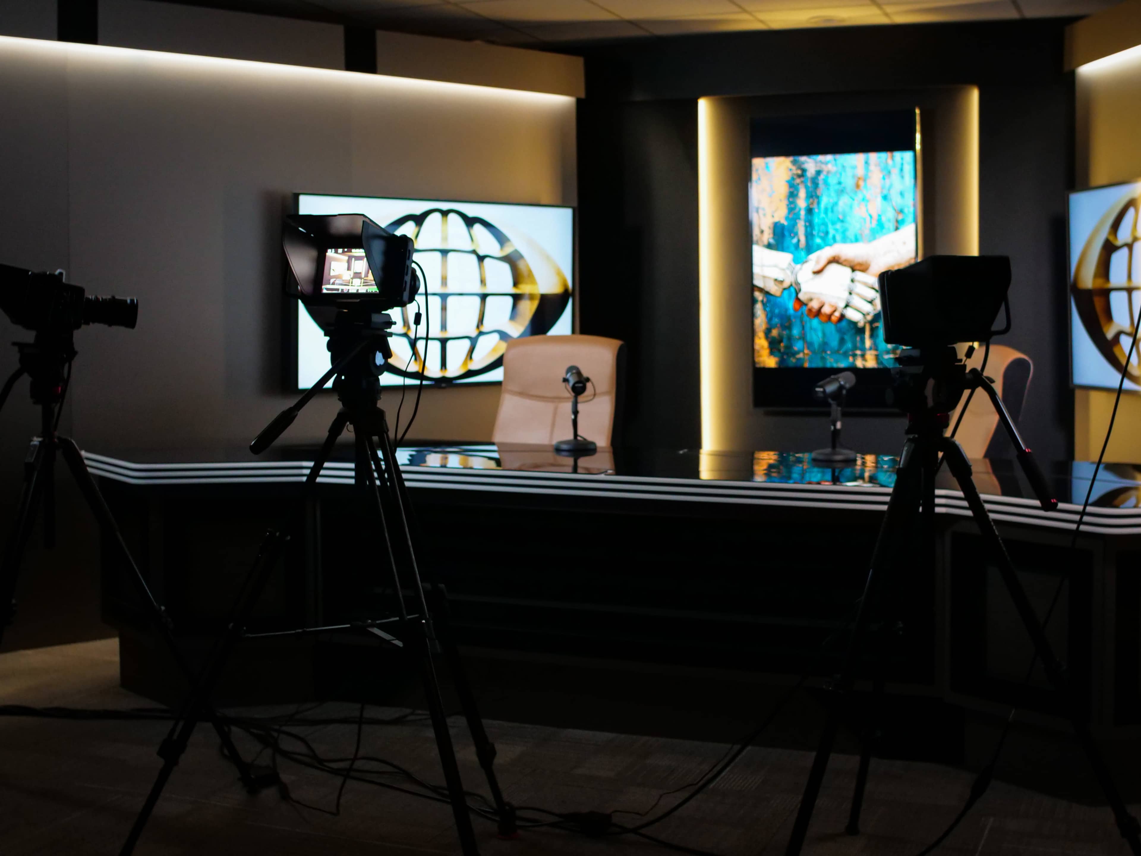 A television studio setup with two cameras positioned on tripods facing a desk, flanked by two screens displaying an artwork of hands shaking.